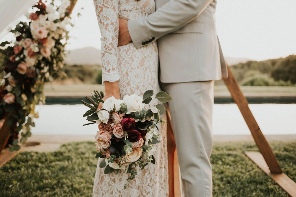 A bride and groom standing close together outdoors, with the bride holding a bouquet of roses and greenery, near a floral arch with mountains and water in the background.