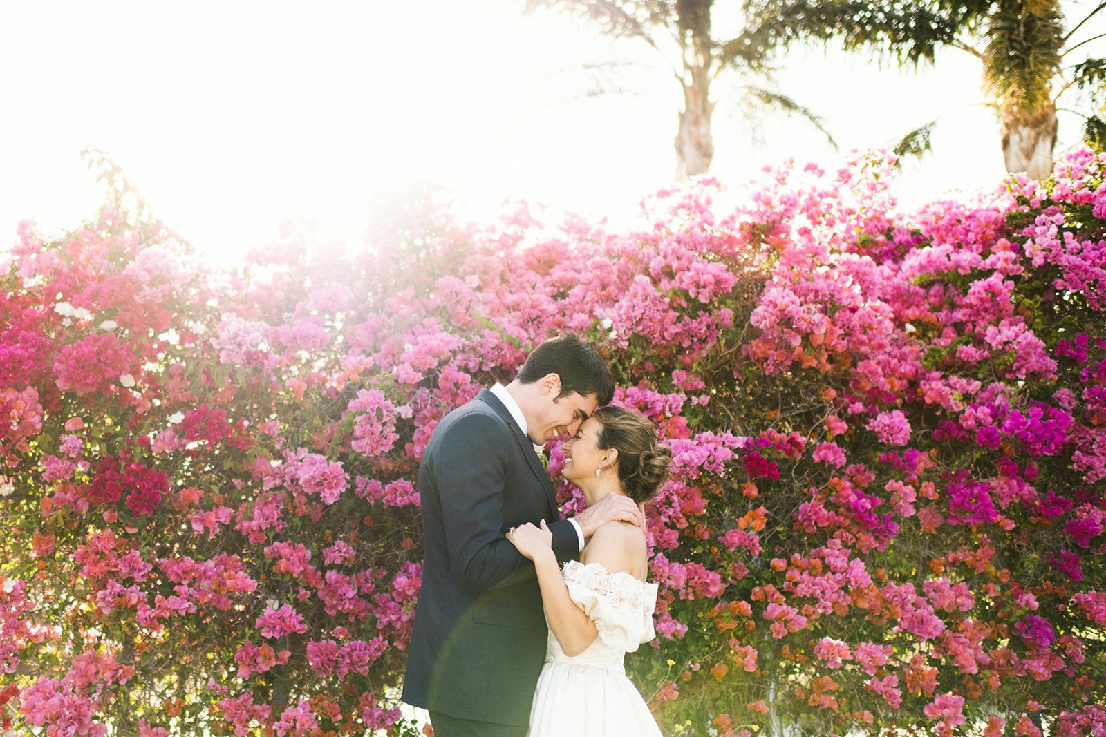 A couple in wedding attire embracing each other in front of pink and purple blooming flowers with palm trees in the background, sunlight shining through.