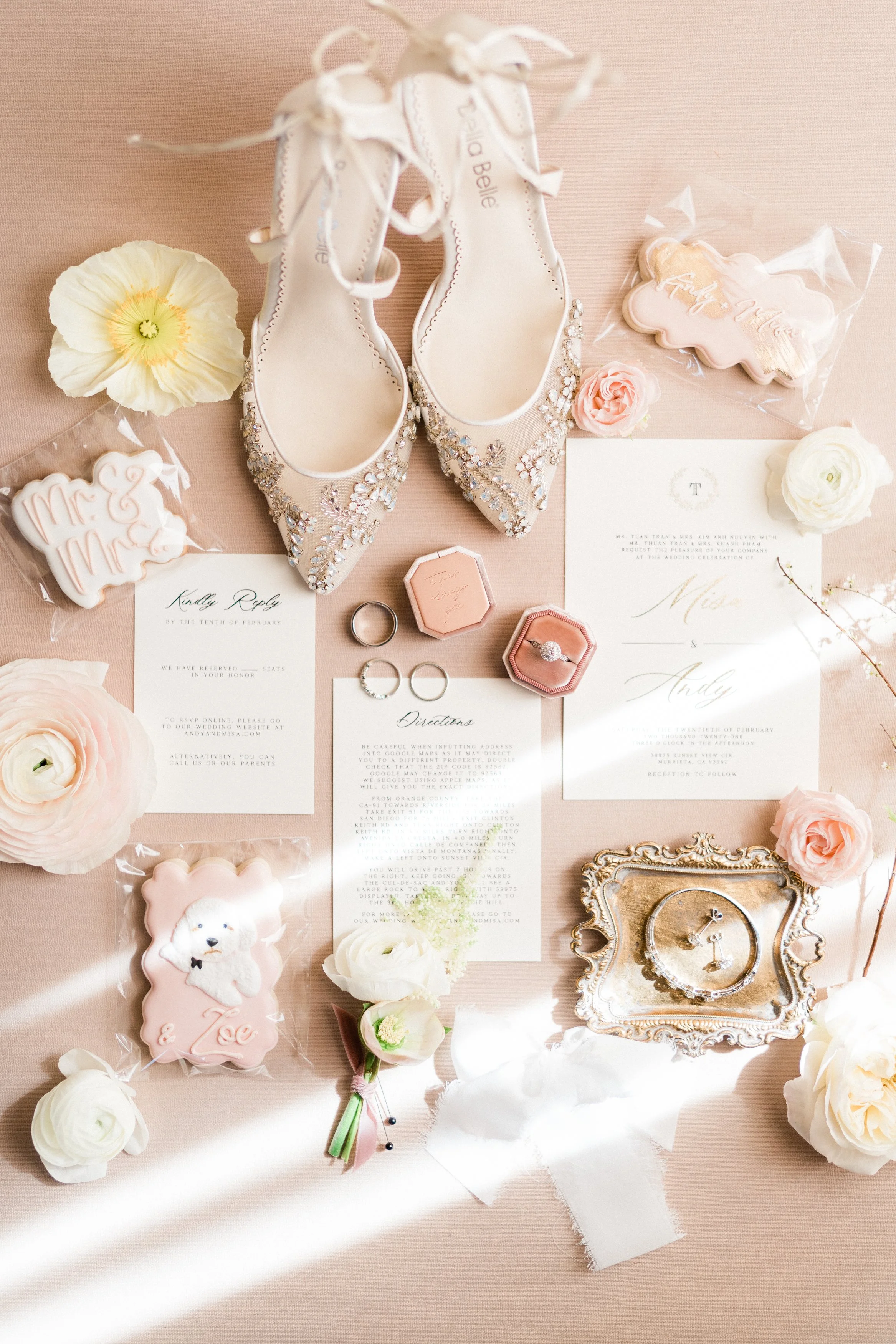 Wedding flat lay with embellished white shoes, wedding invitations, rings, cookies, flowers, and a vintage clock on a pink surface.