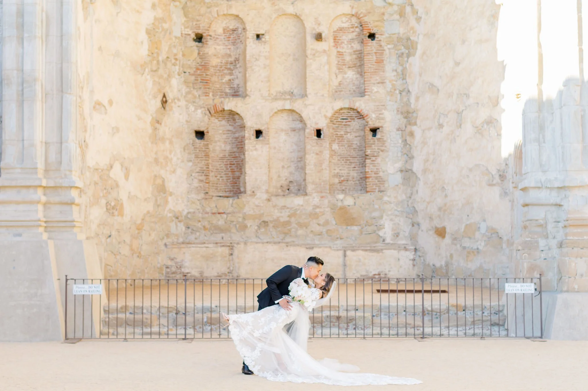 A newlywed couple, dressed in a wedding gown and tuxedo, share a kiss in front of an ancient stone and brick wall with arches and small niches.