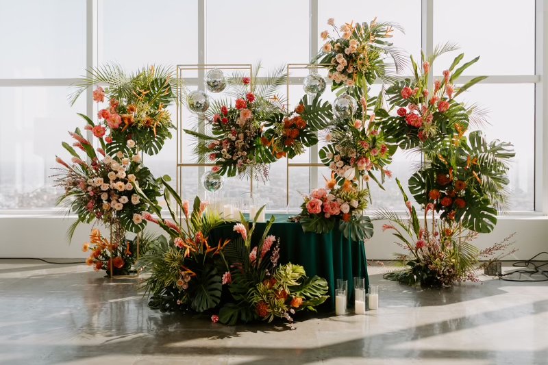A vibrant floral display with pink, orange, and white flowers, green tropical leaves, decorative glass spheres, and candles in a bright, sunlit room with large windows.