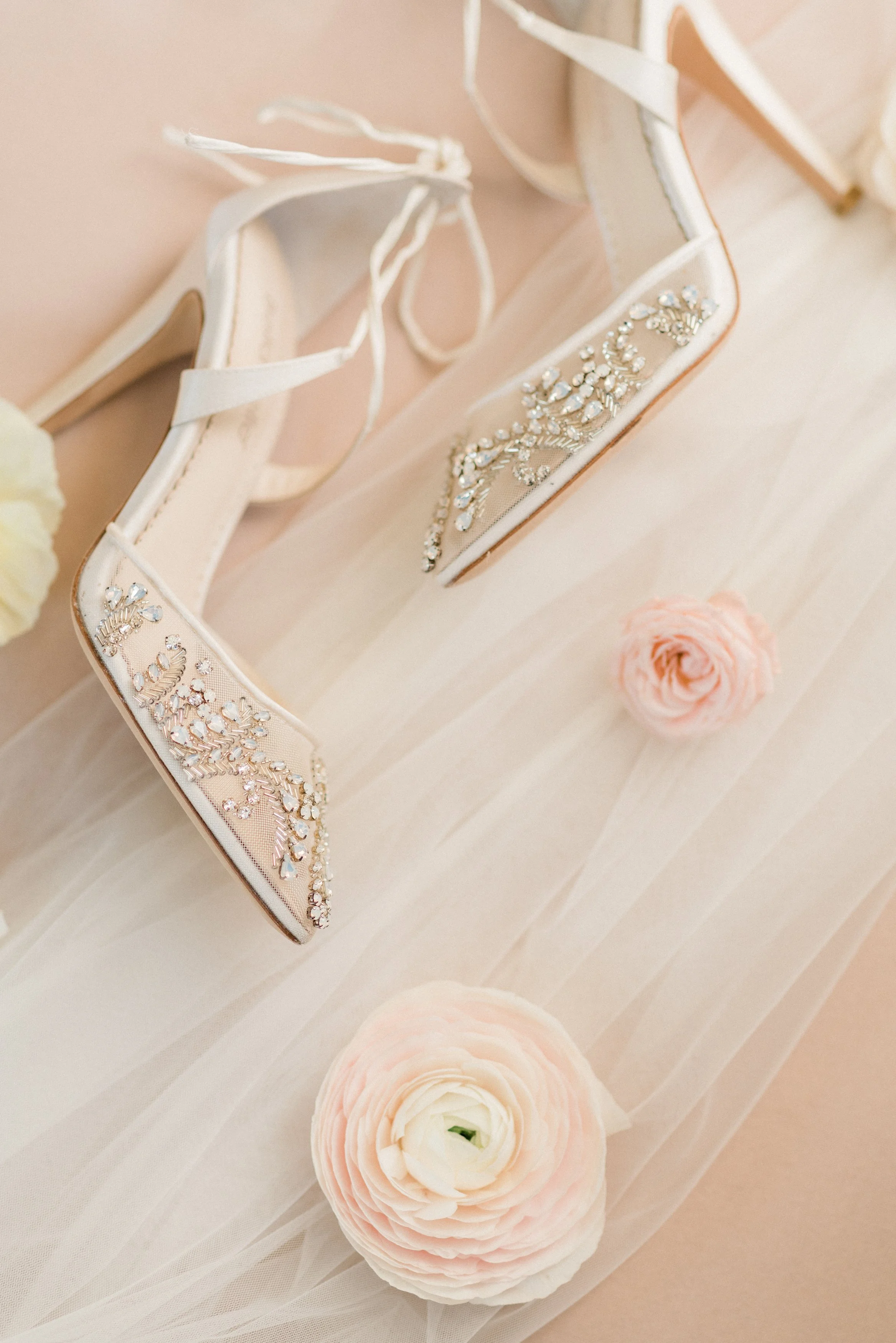 A pair of white high-heeled shoes with rhinestone embellishments, surrounded by pink and white flowers, set on a light pink and cream-colored background.