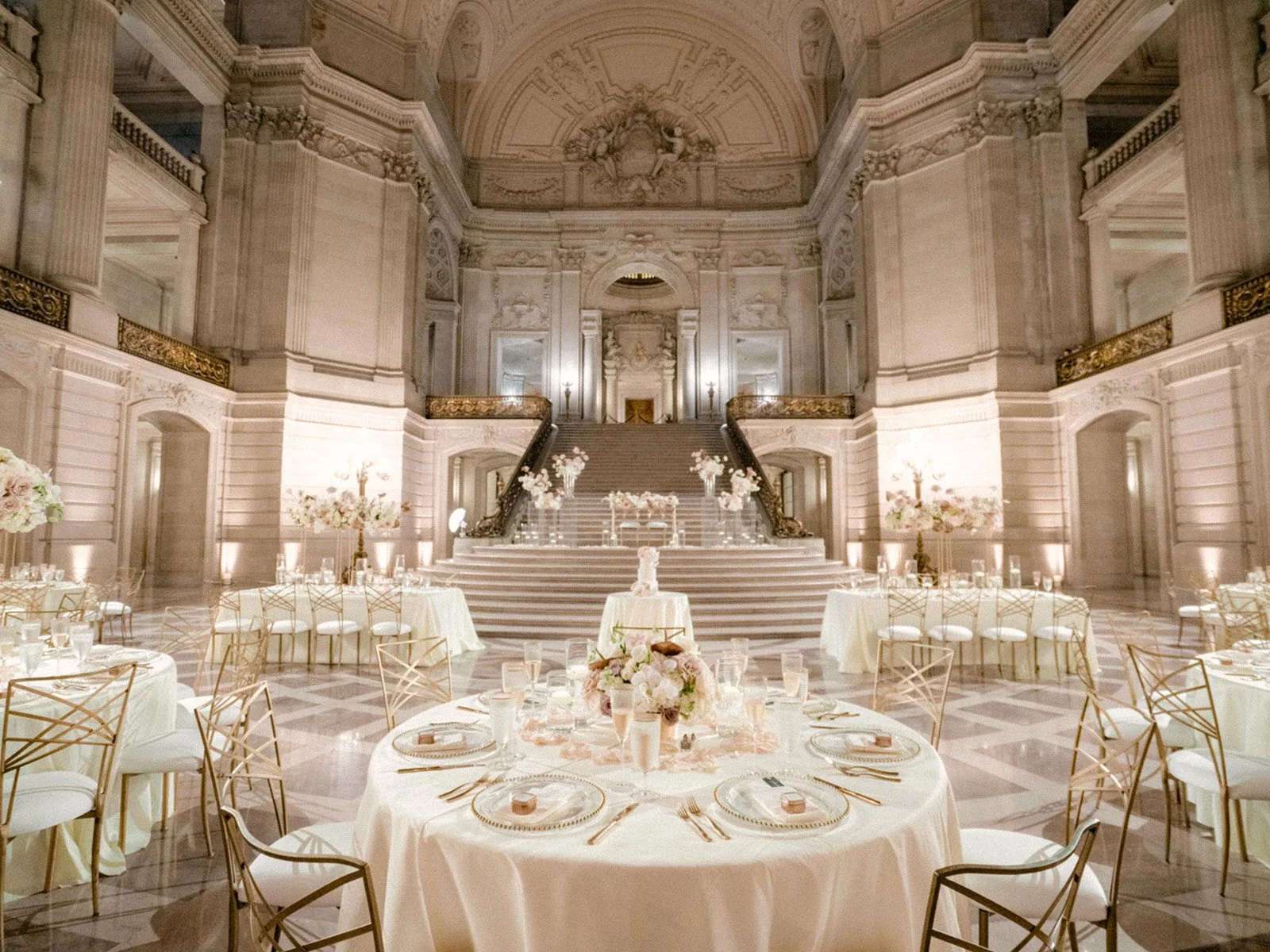 Elegant banquet hall decorated for an event with round tables with white tablecloths, gold chairs, floral centerpieces, and a staircase with floral arrangements leading up to a stage.