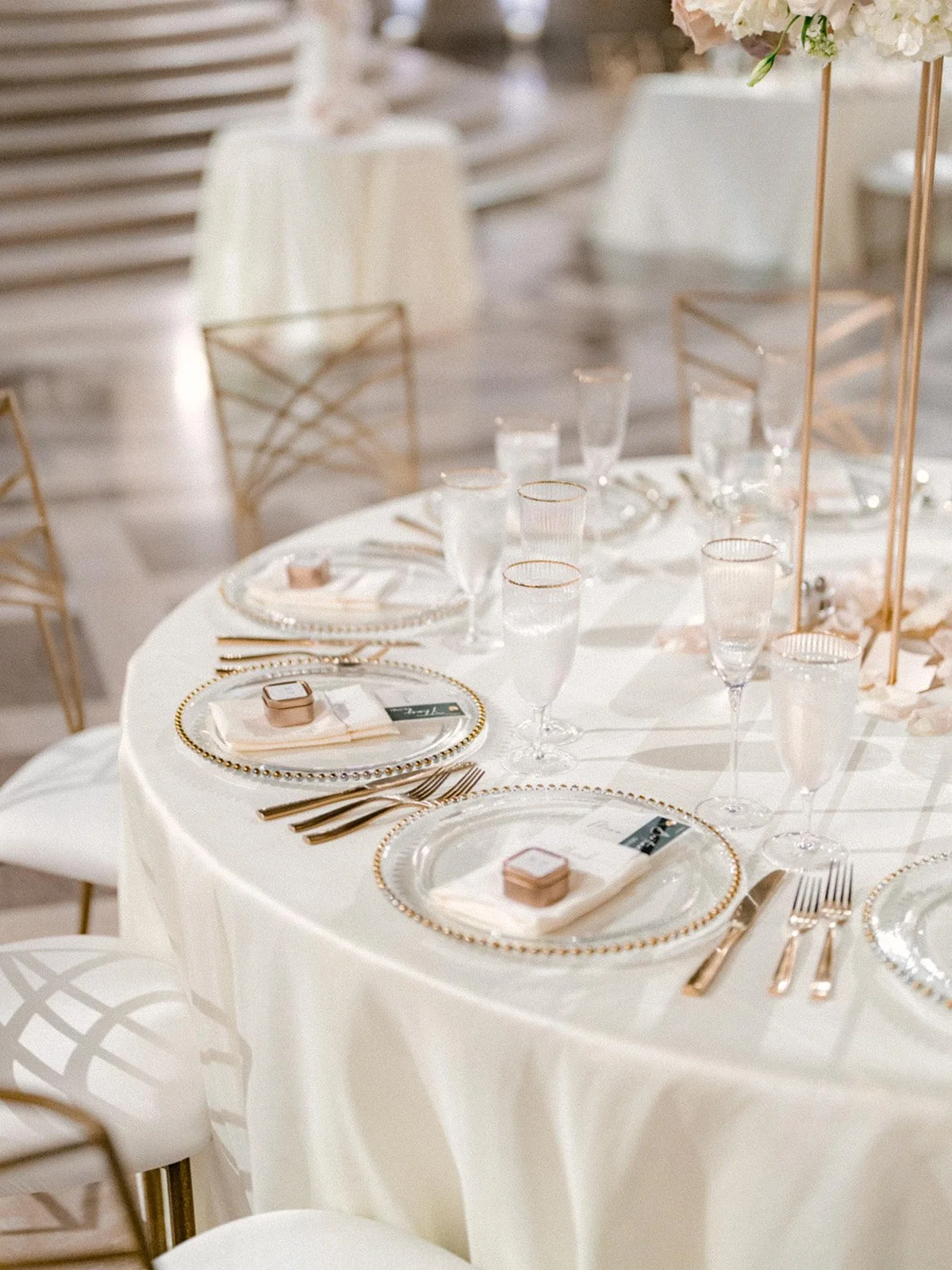 Elegant round table set for a formal event with white tablecloth, clear glass plates with gold beaded rim, gold utensils, wine glasses, small favor boxes on plates, and a tall floral centerpiece with white blooms.