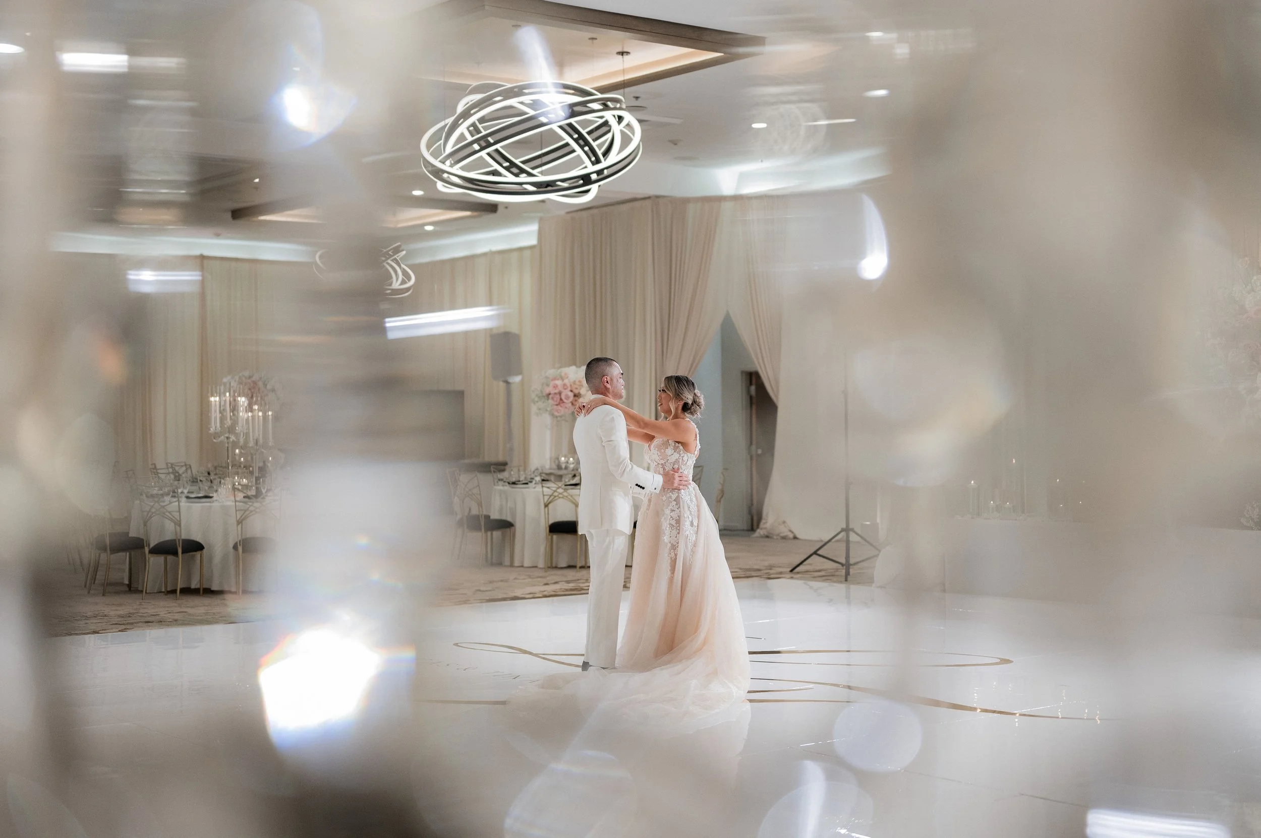 A bride and groom dancing in a wedding reception hall, viewed through a decorative mirror or glass with a blurred, sparkly effect.