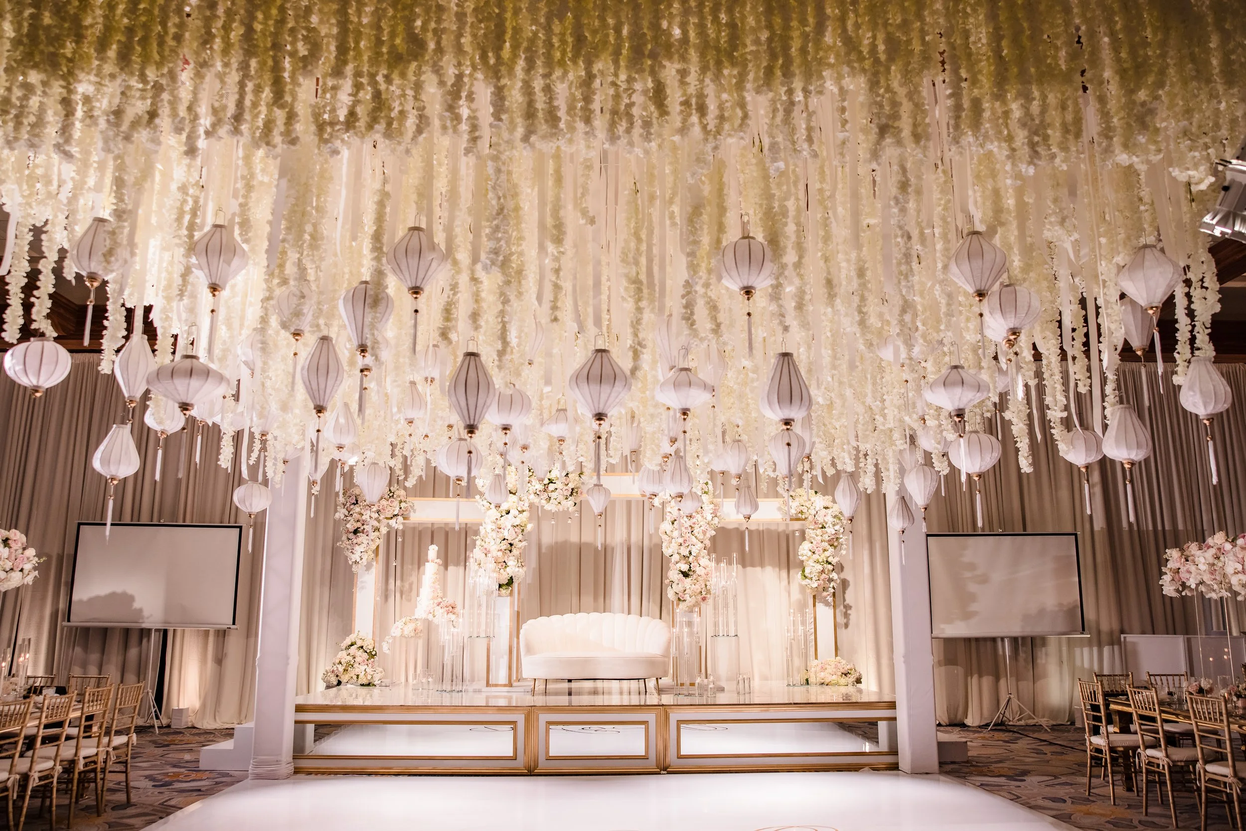 Elegant wedding stage decorated with hanging white lanterns and flowers, featuring a white sofa in the center, with floral arrangements and projection screens on sides.
