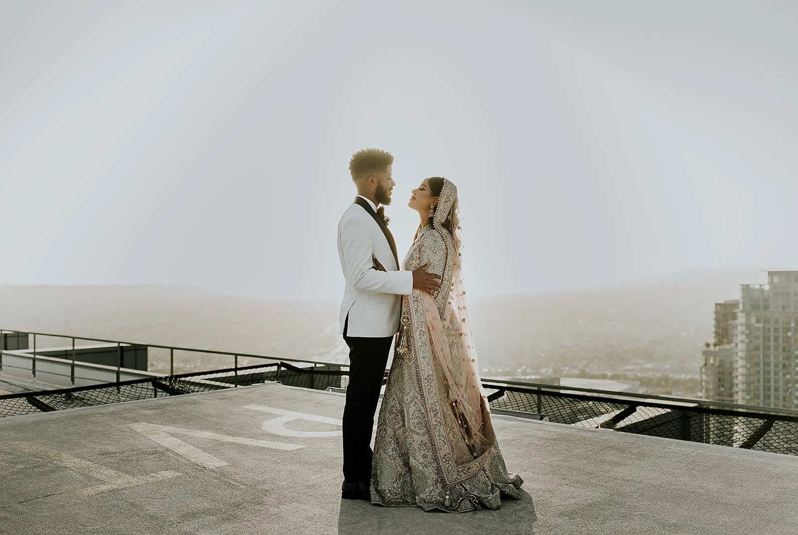 A couple dressed in wedding attire standing on a rooftop, looking into each other's eyes, with a city skyline in the background.
