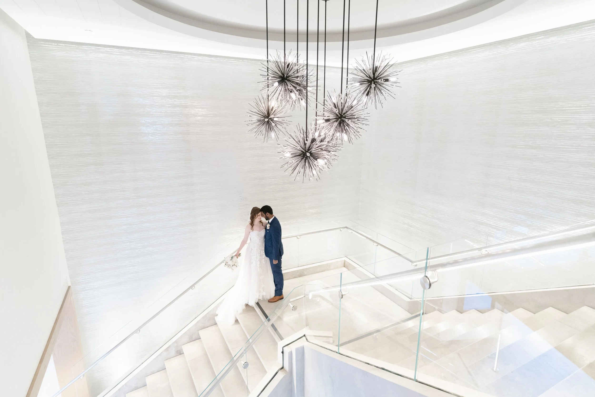 A bride and groom standing on a staircase at their wedding, touching foreheads, with a modern chandelier hanging above them and a white textured wall in the background.