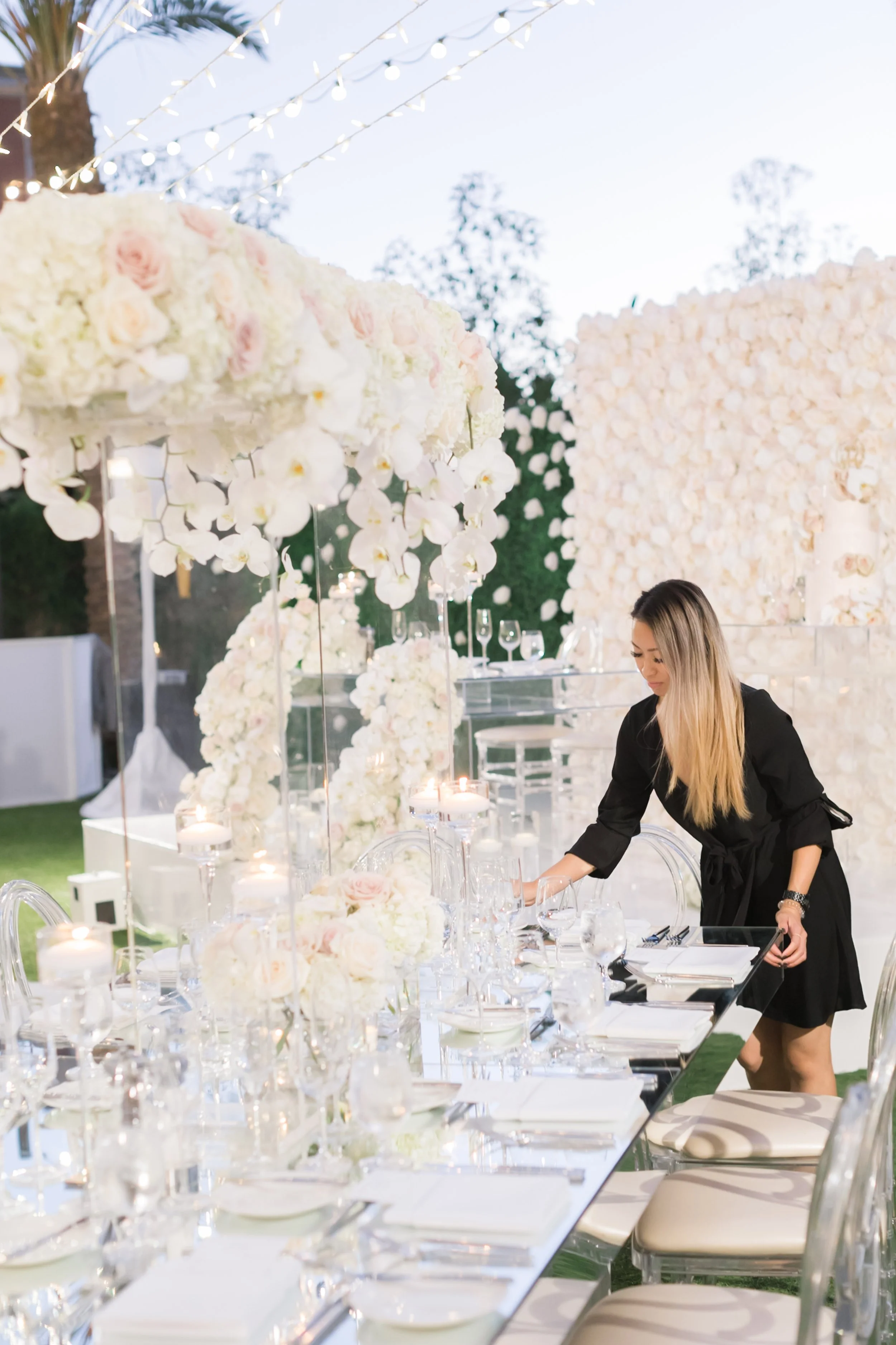 A woman setting a table for a wedding reception outdoors, decorated with white floral arrangements, candles, and glassware during dusk.