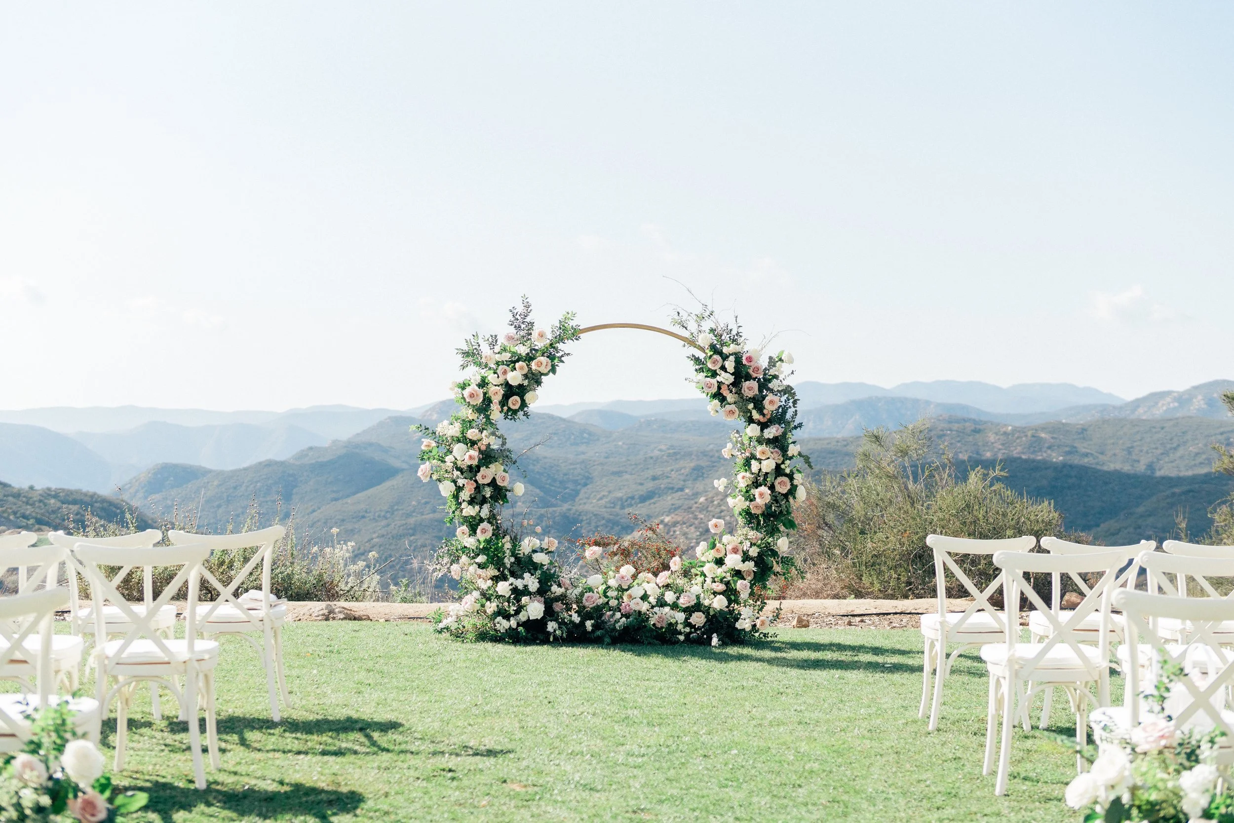 Wedding altar with floral arch and chairs on grass, mountain scenery in the background.