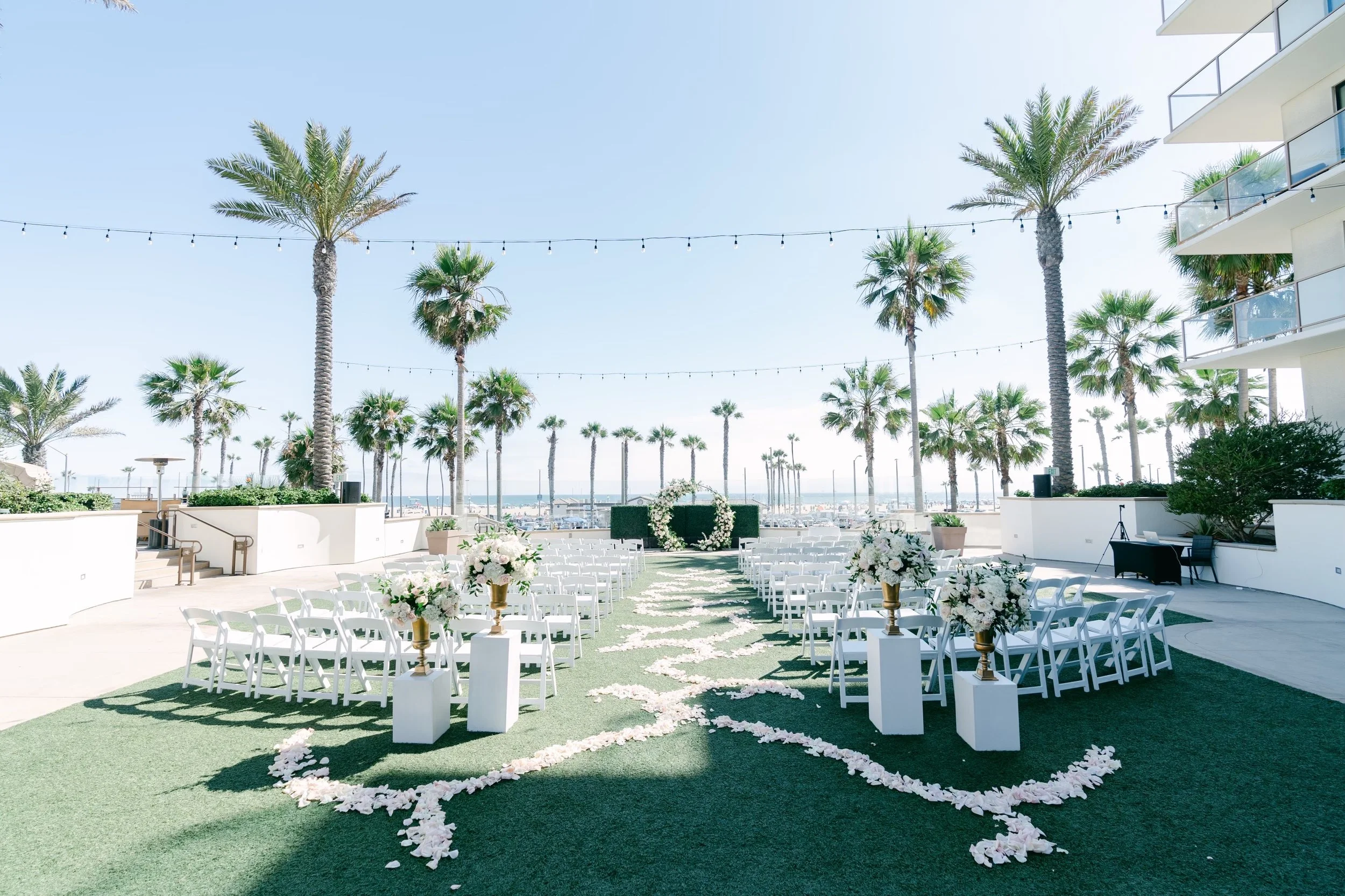Outdoor wedding setup with white chairs, floral arrangements, and a heart-shaped flower arch on a grassy aisle, under string lights, overlooking a marina with palm trees and a clear sky.