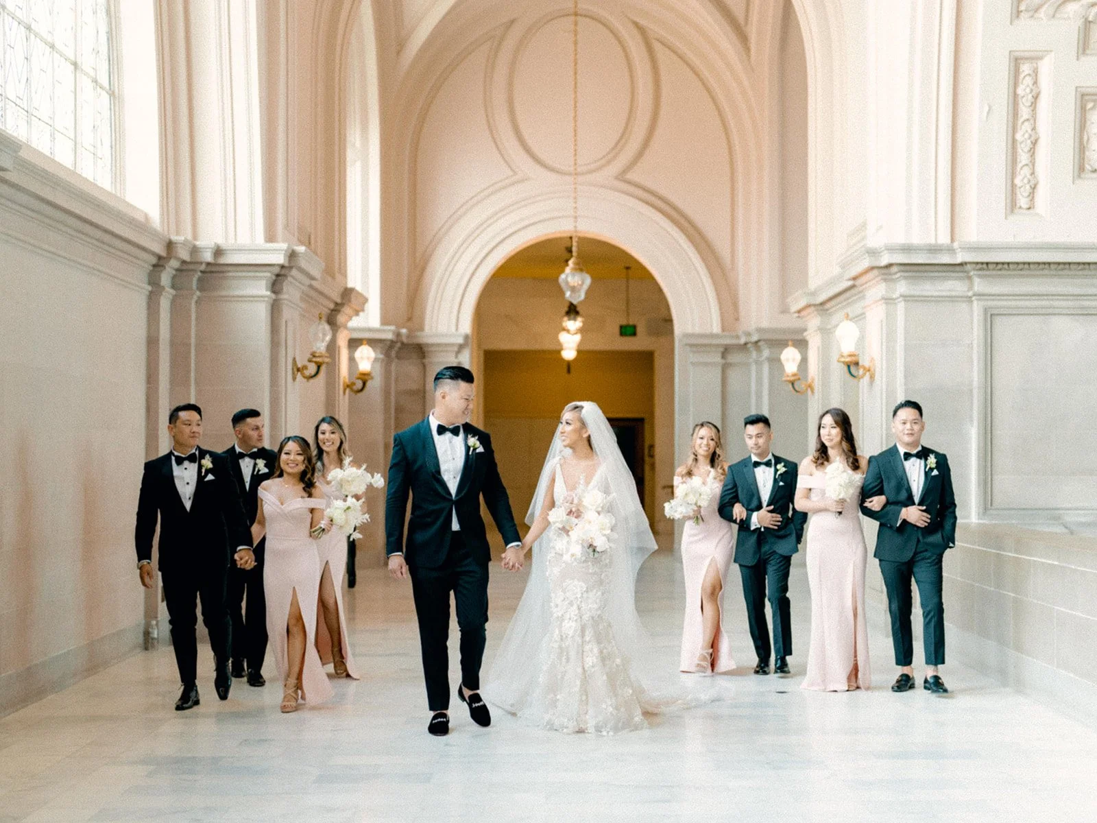Bride and groom walking hand in hand inside a grand hall with wedding party.