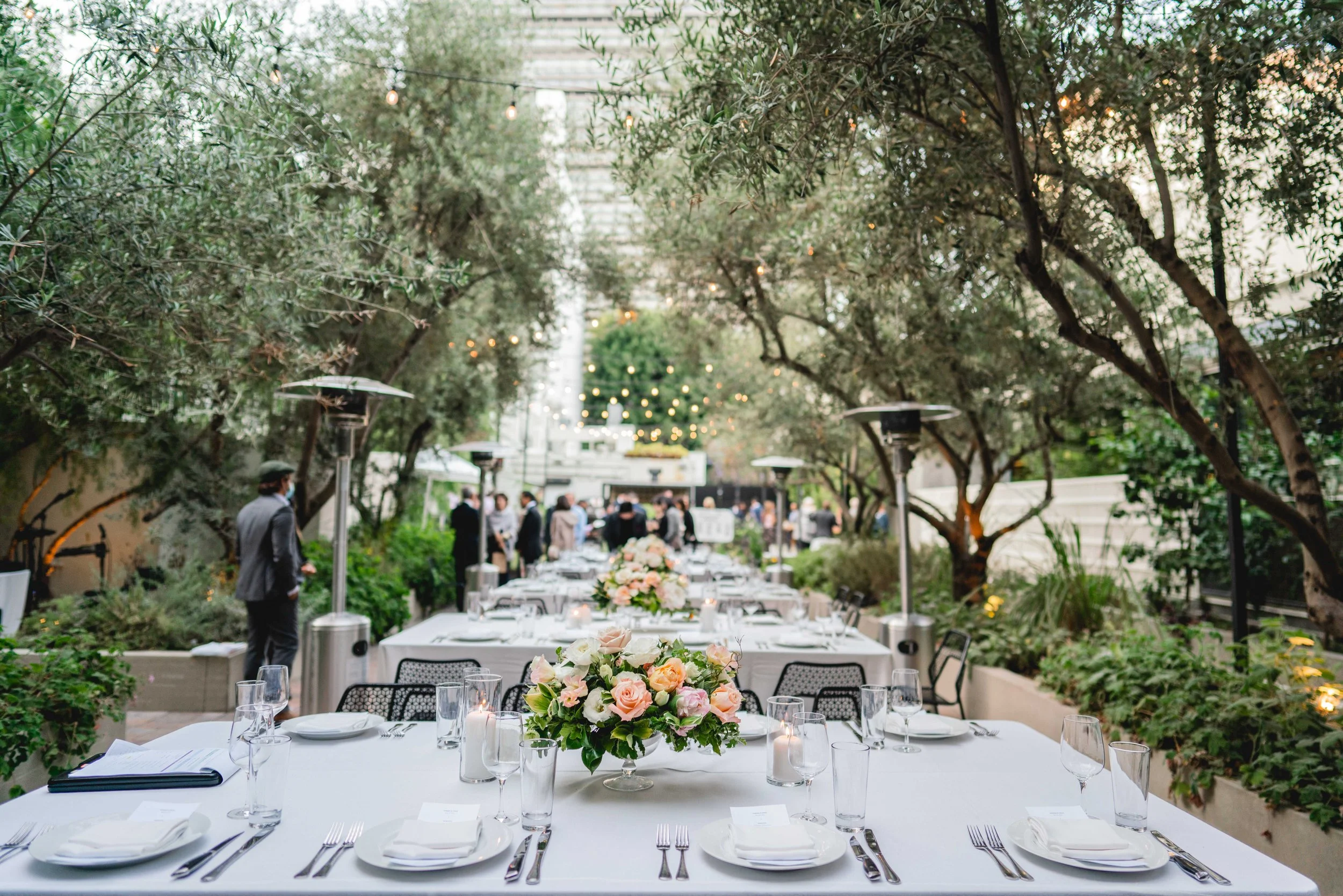 Outdoor banquet table set with white tablecloths, floral centerpieces, glassware, and plates, under string lights and surrounded by trees.