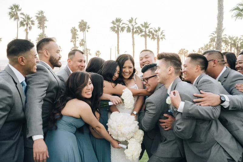 Happy wedding party group of men and women in formal attire, posing and smiling outdoors with palm trees in the background.
