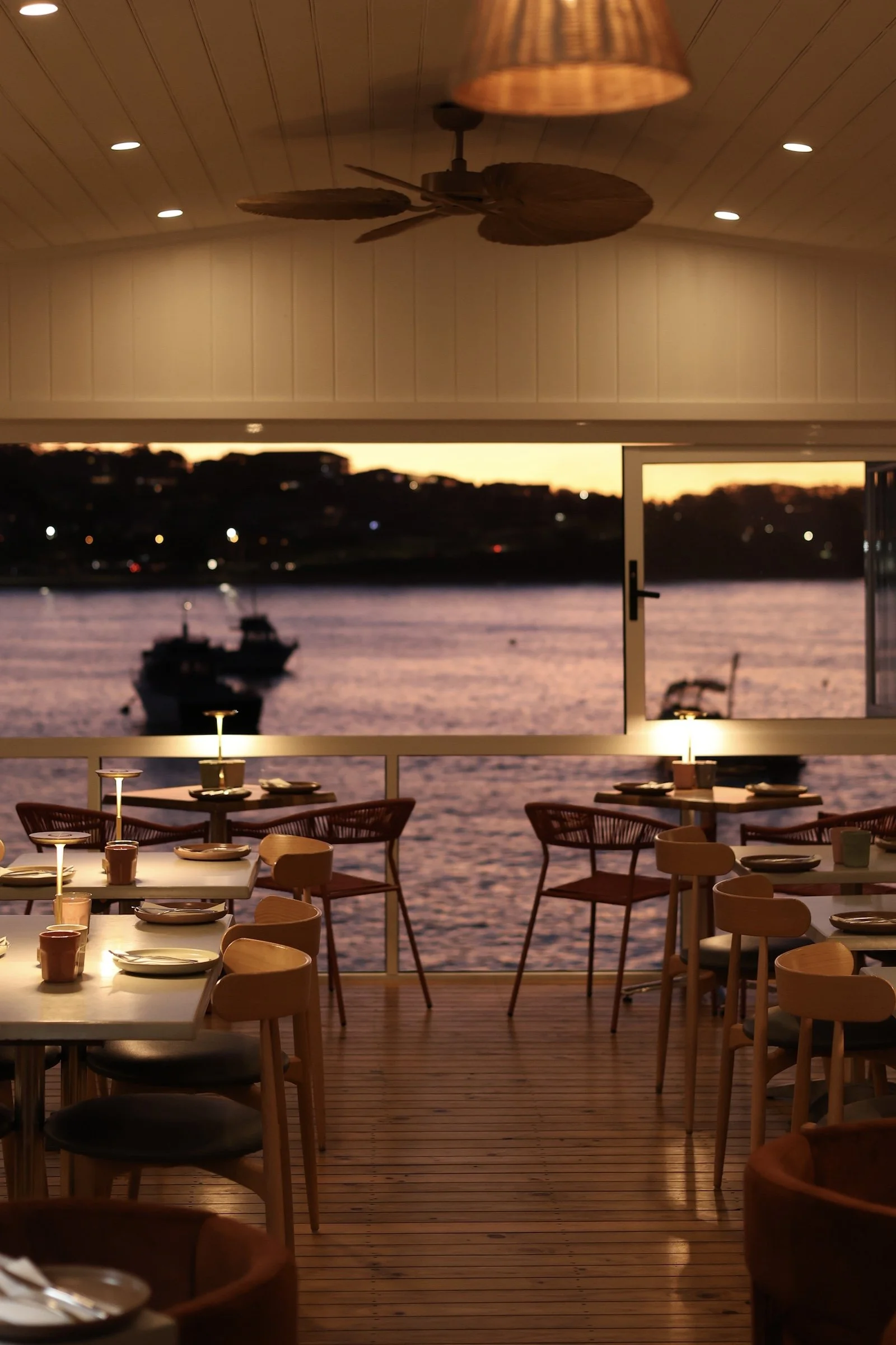 Interior of a restaurant with tables set for dining, overlooking water and boats during sunset.