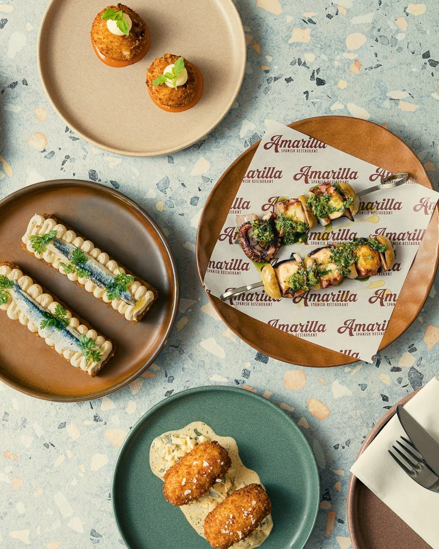 Assorted Spanish dishes on colorful plates, including croquettes with sauce, grilled octopus skewers, green sprigs on seafood, and a garnished dessert with cream, presented on a terrazzo tabletop.
