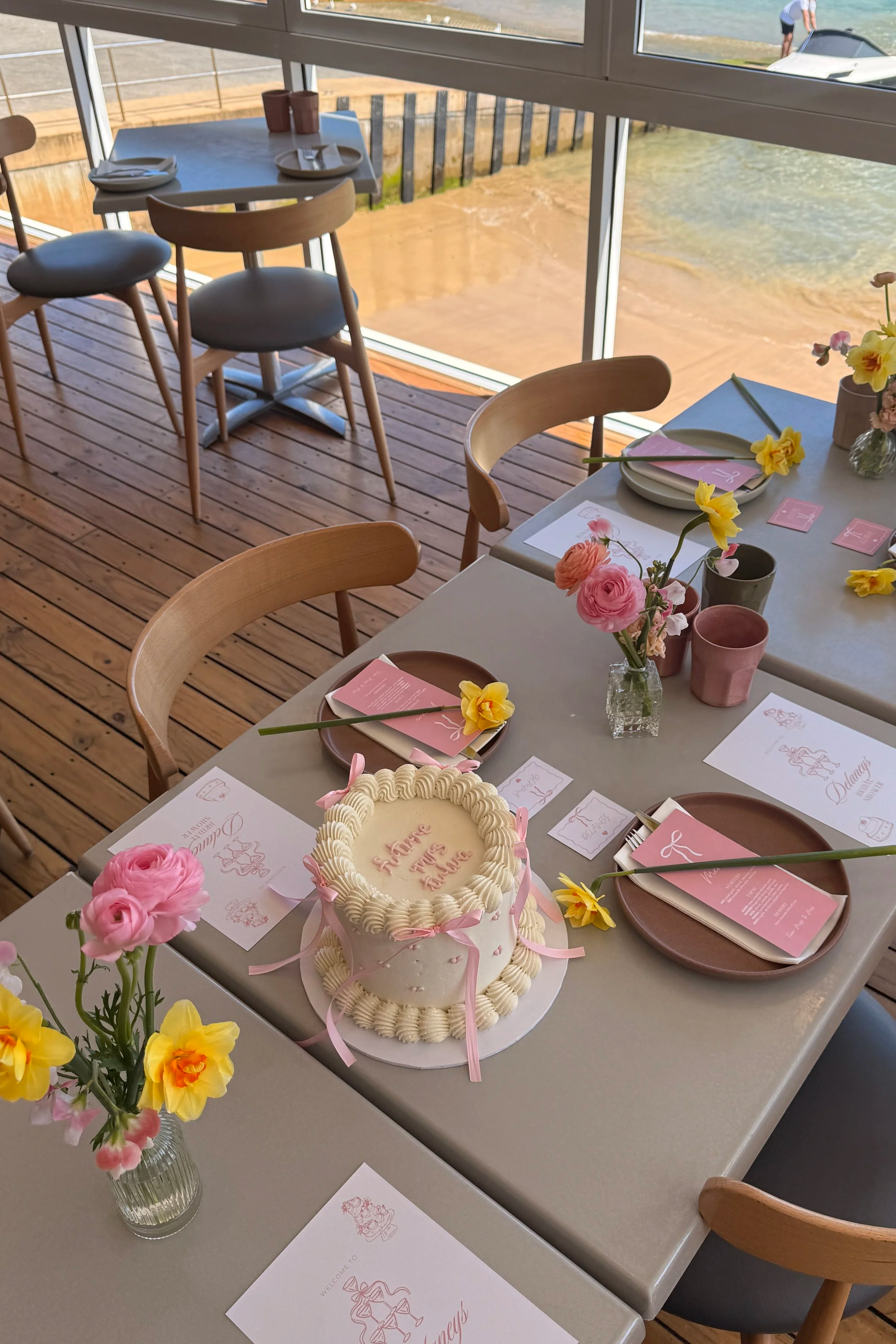 A table set for a celebration with a pink and white cake, pink and yellow flowers in vases, pink and brown plates with pink napkins and cutlery, and printed menus or invitations. The setting overlooks a body of water outside a window.