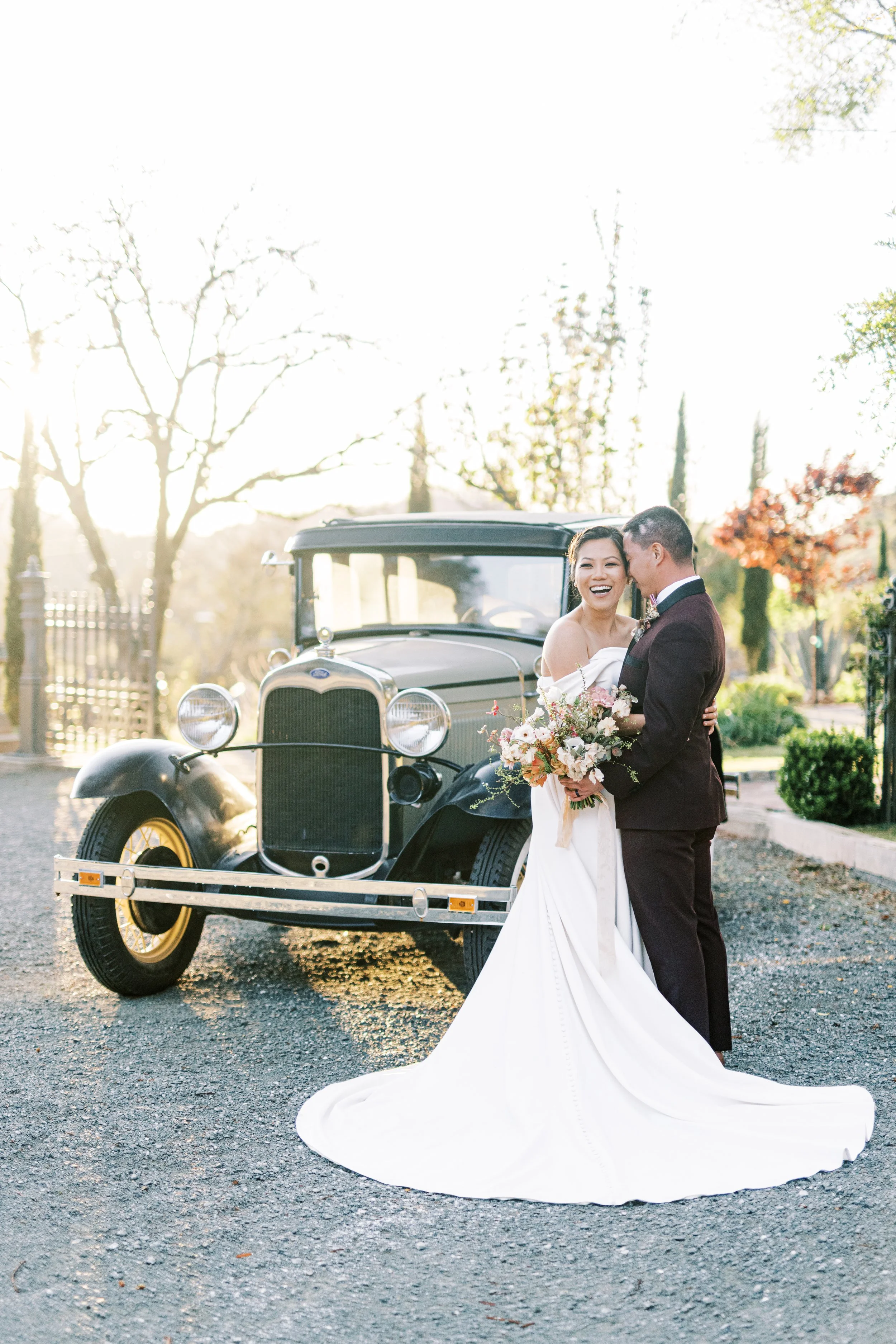 Bride and groom embracing in front of a vintage black car with yellow wheels under the trees at Triple S Ranch Napa.