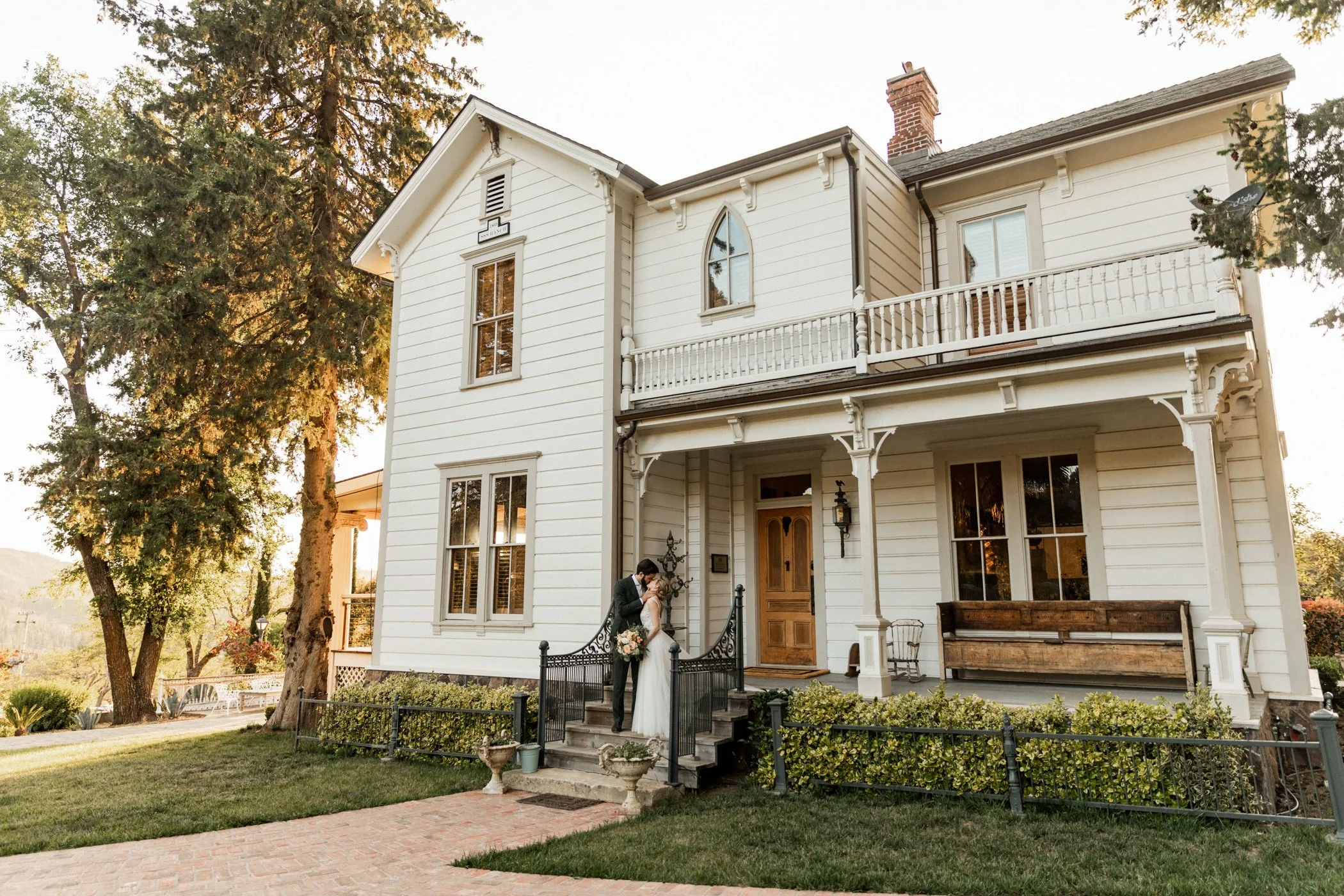 Couple in wedding attire on the front steps of a two-story white house with a porch and trees around.