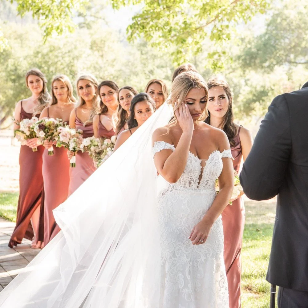 Bride touching her temple during ceremony, bridesmaids with bouquets standing behind her in green outdoor setting.