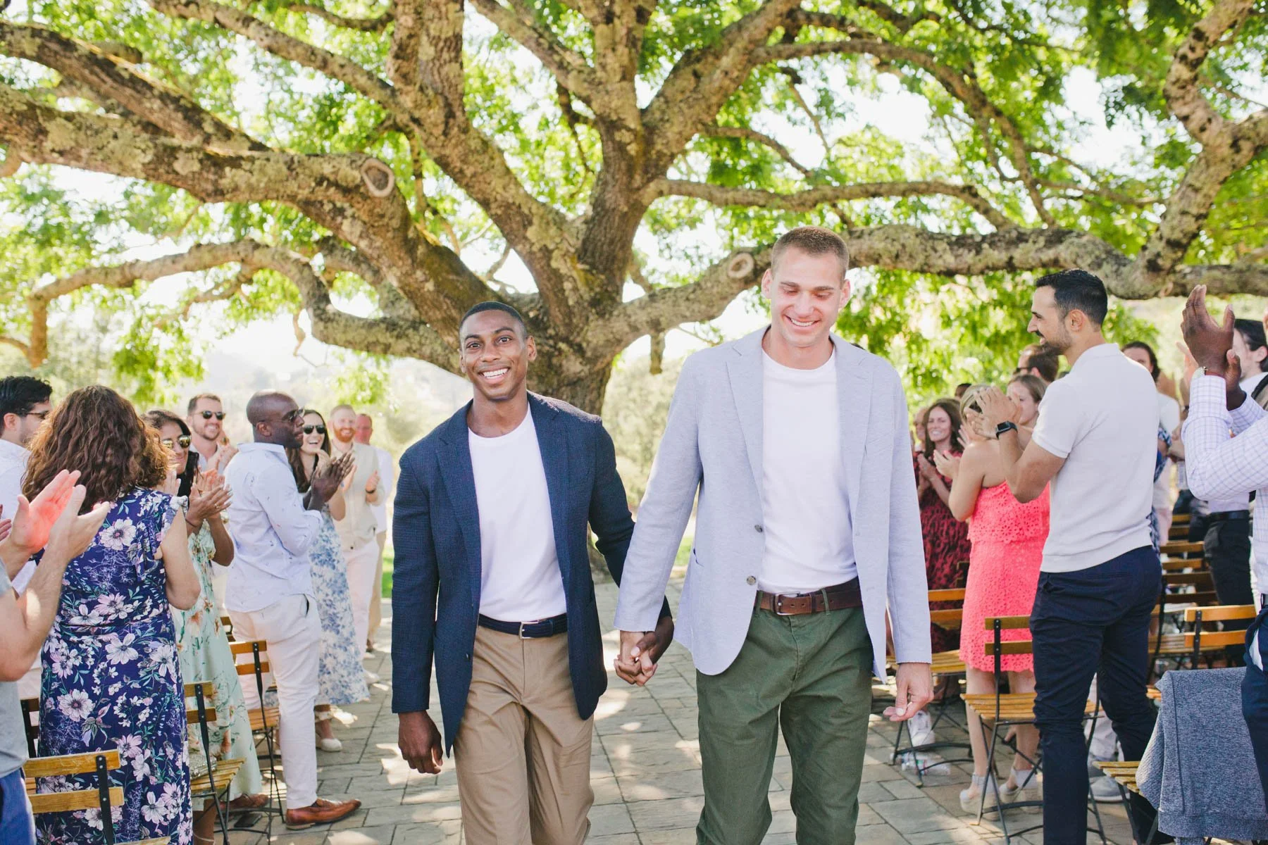 Two grooms walking down the aisle hand in hand at outdoor wedding under a large tree, surrounded by cheering guests.