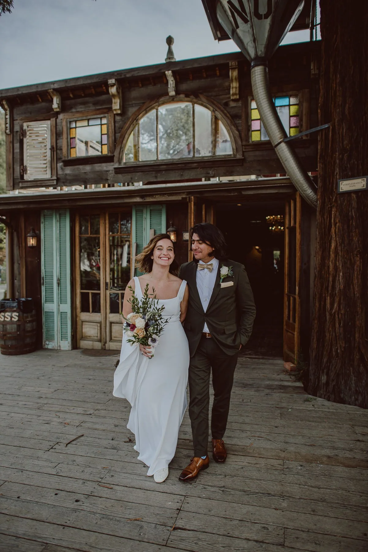Bride and groom walking outside rustic wooden building on wedding day, smiling and holding bouquet.
