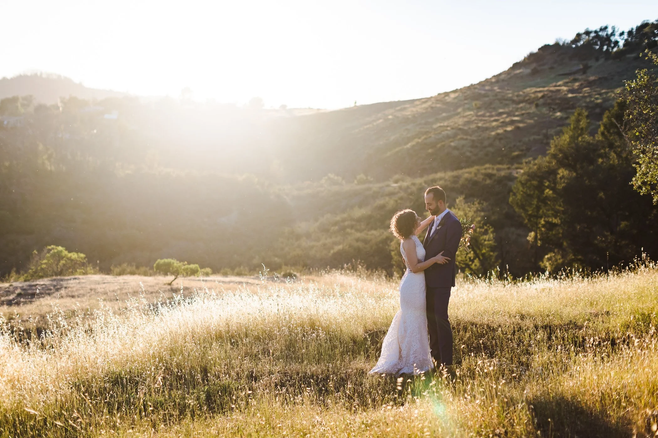 Couple standing together in a grassy field at sunset, embracing with rolling hills and trees in the background.