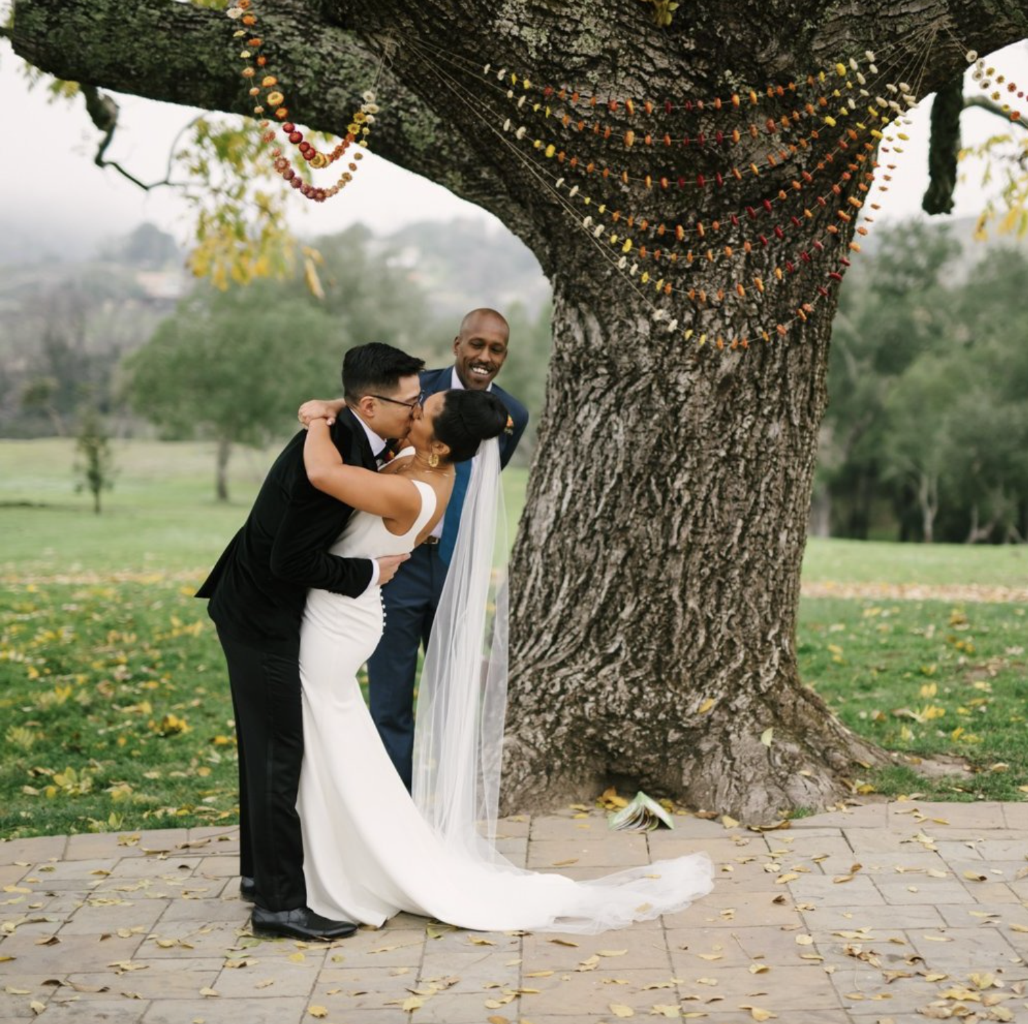 Wedding couple kissing beneath large tree decorated with colorful hanging beads, officiant smiling nearby.