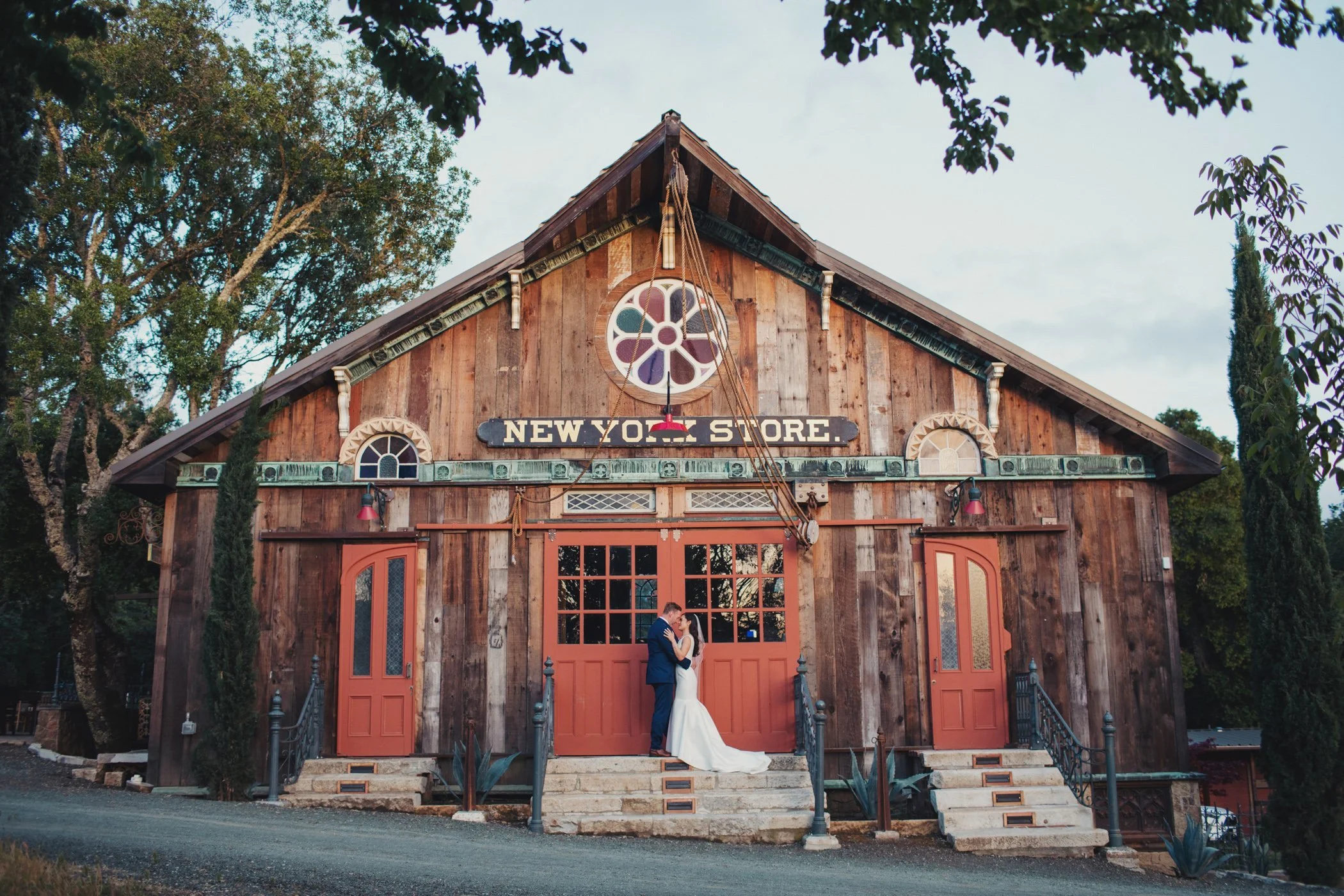A wedding couple embraces in front of a rustic wooden building labeled 'New York Store', with steps leading up to red doors and surrounding greenery.