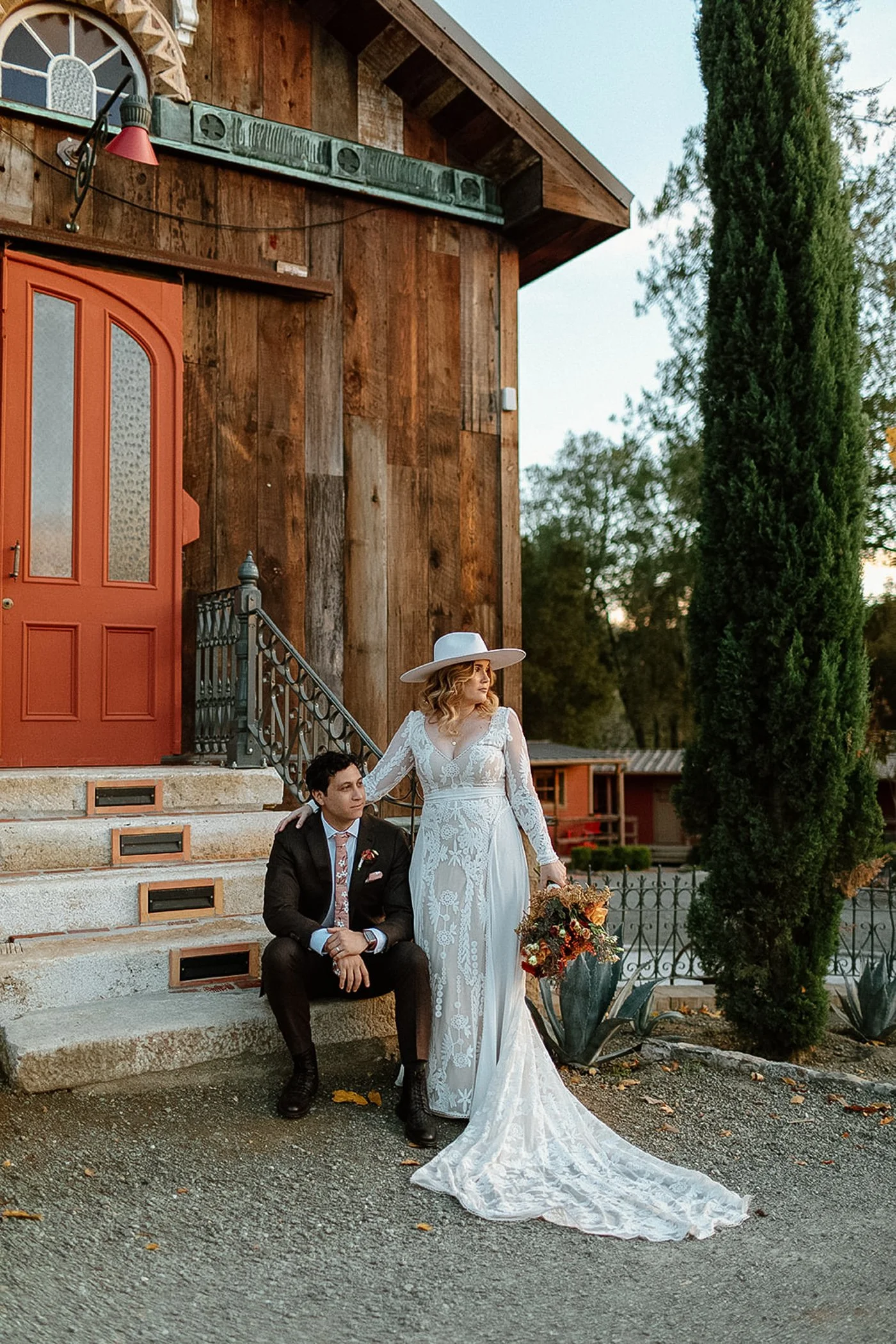 Bride in lace dress and wide-brimmed hat standing with groom in colorful tie outside rustic red-doored building.
