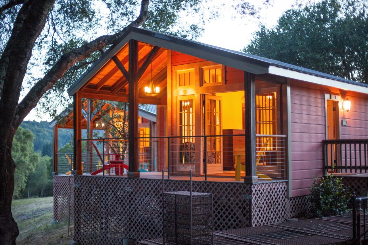A cozy wooden house with a screened porch, lit from inside, surrounded by trees and greenery.