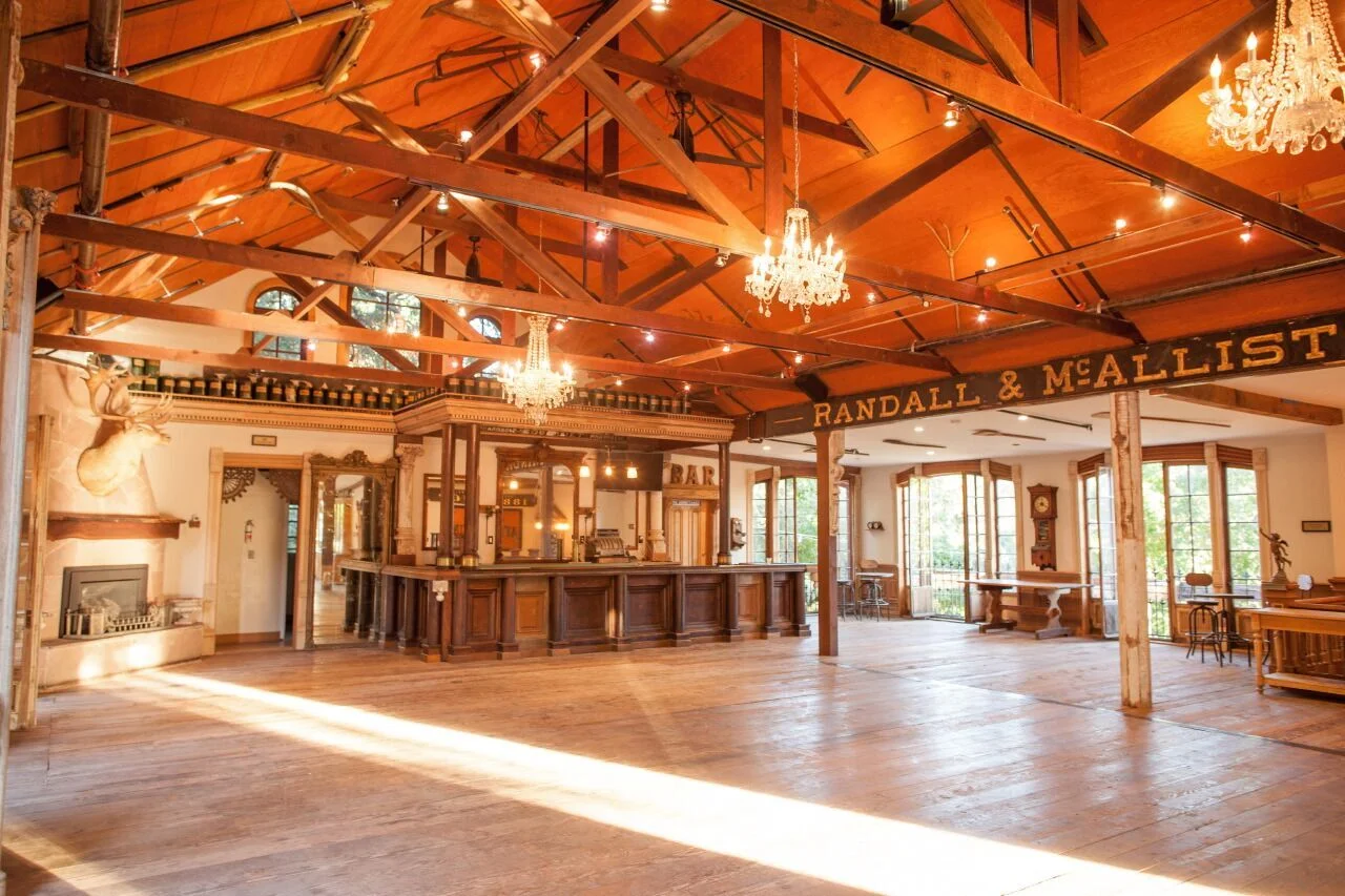 Interior of a rustic event space with wooden floors, exposed wooden beams, chandeliers, and large windows, featuring a bar labeled 'RANDLE & MALLIST'.