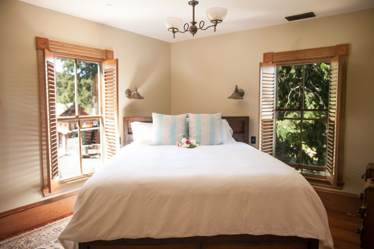 A cozy bedroom with a large bed, white bedding, and striped pillows. Two wooden windows with shutters provide views of trees outside. A chandelier hangs from the ceiling, and small wall-mounted lamps are on either side of the bed.