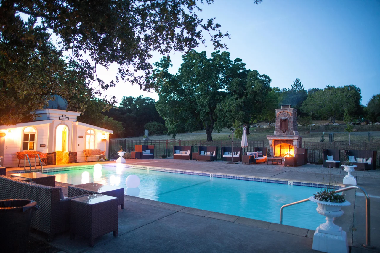 Outdoor swimming pool area at dusk with lounge chairs, a fireplace, and surrounding trees.