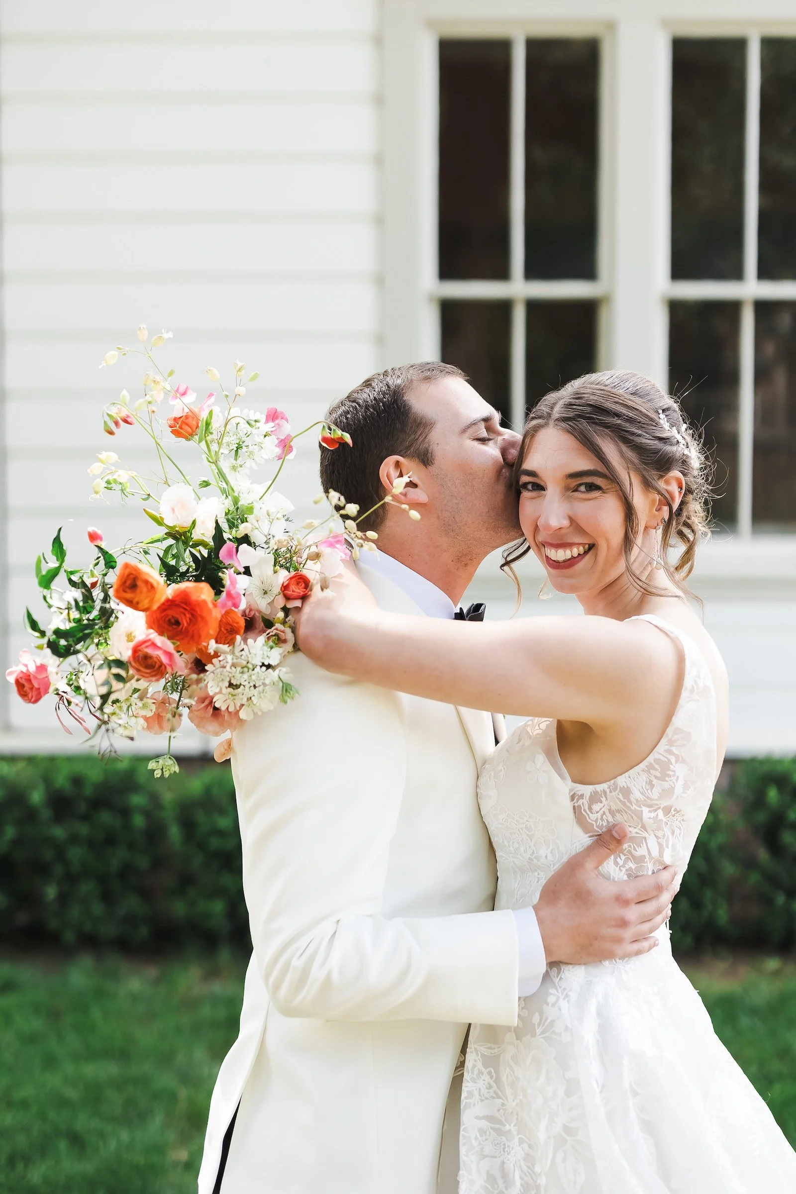 Newlywed couple embracing outdoors, groom kissing bride’s cheek as she smiles and holds a colorful bouquet.