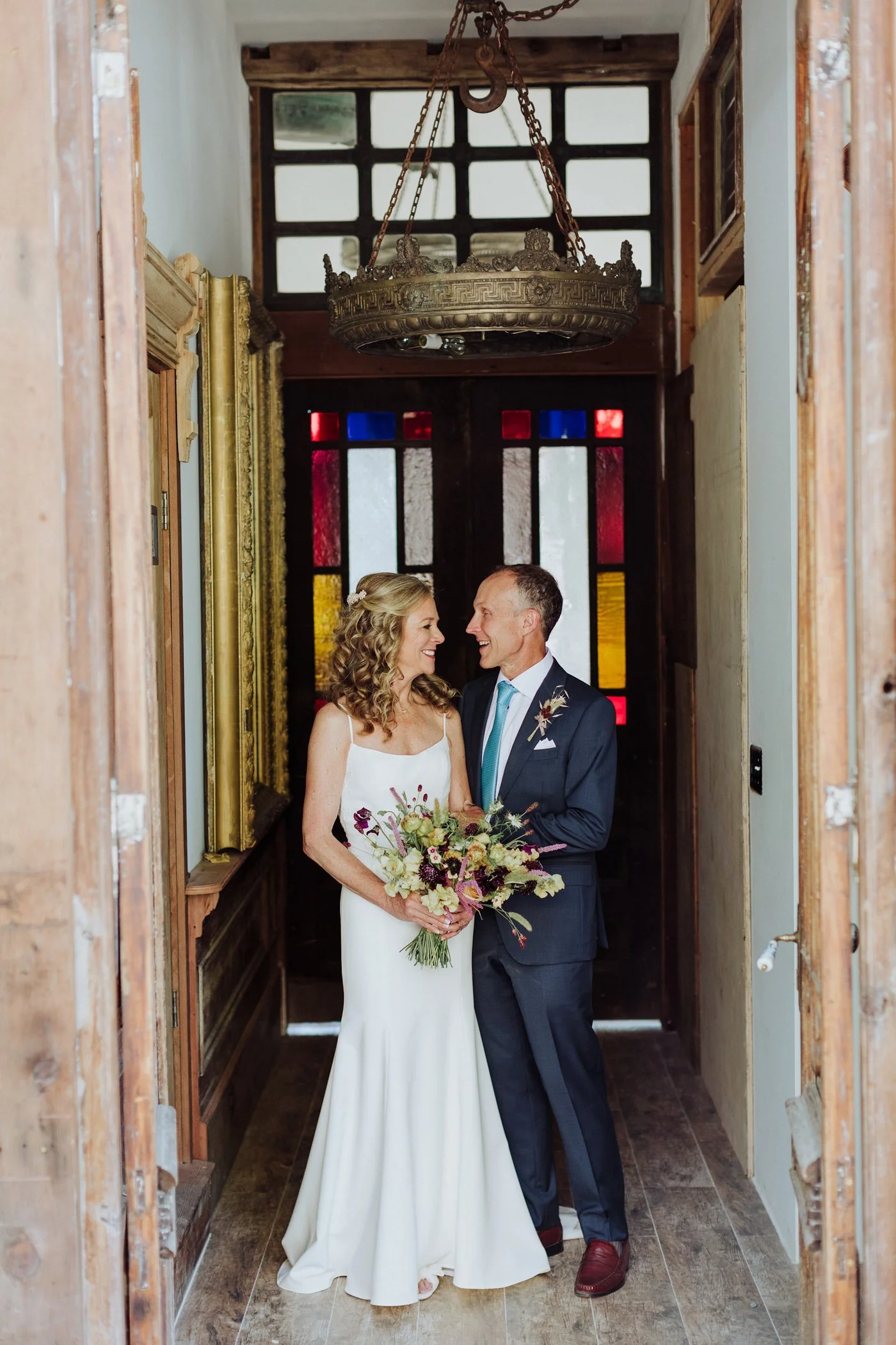 Couple standing in wooden hallway with stained glass windows, laughing together with bouquet in hand