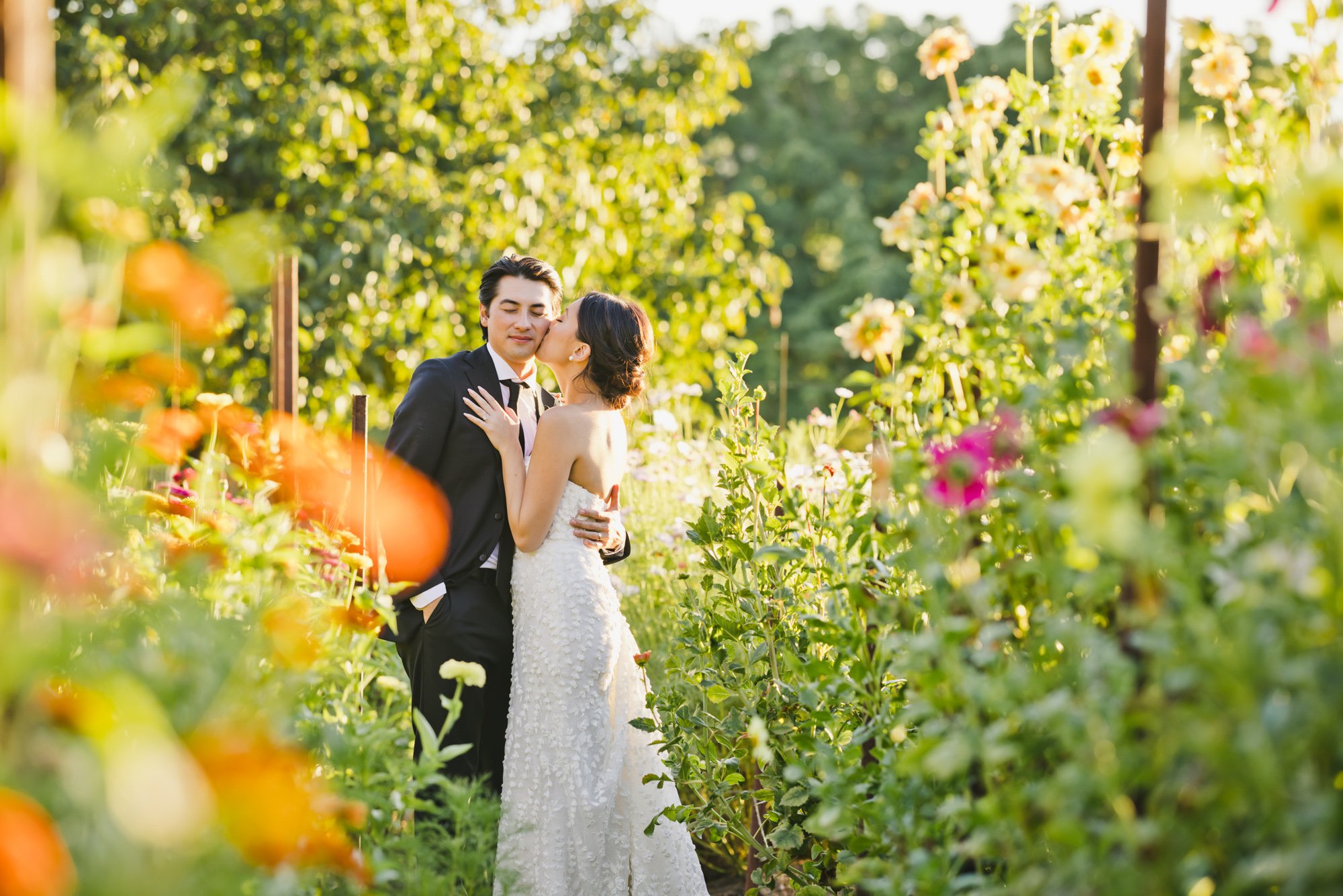 Bride and groom kissing in a colorful garden surrounded by flowers and trees during golden hour.