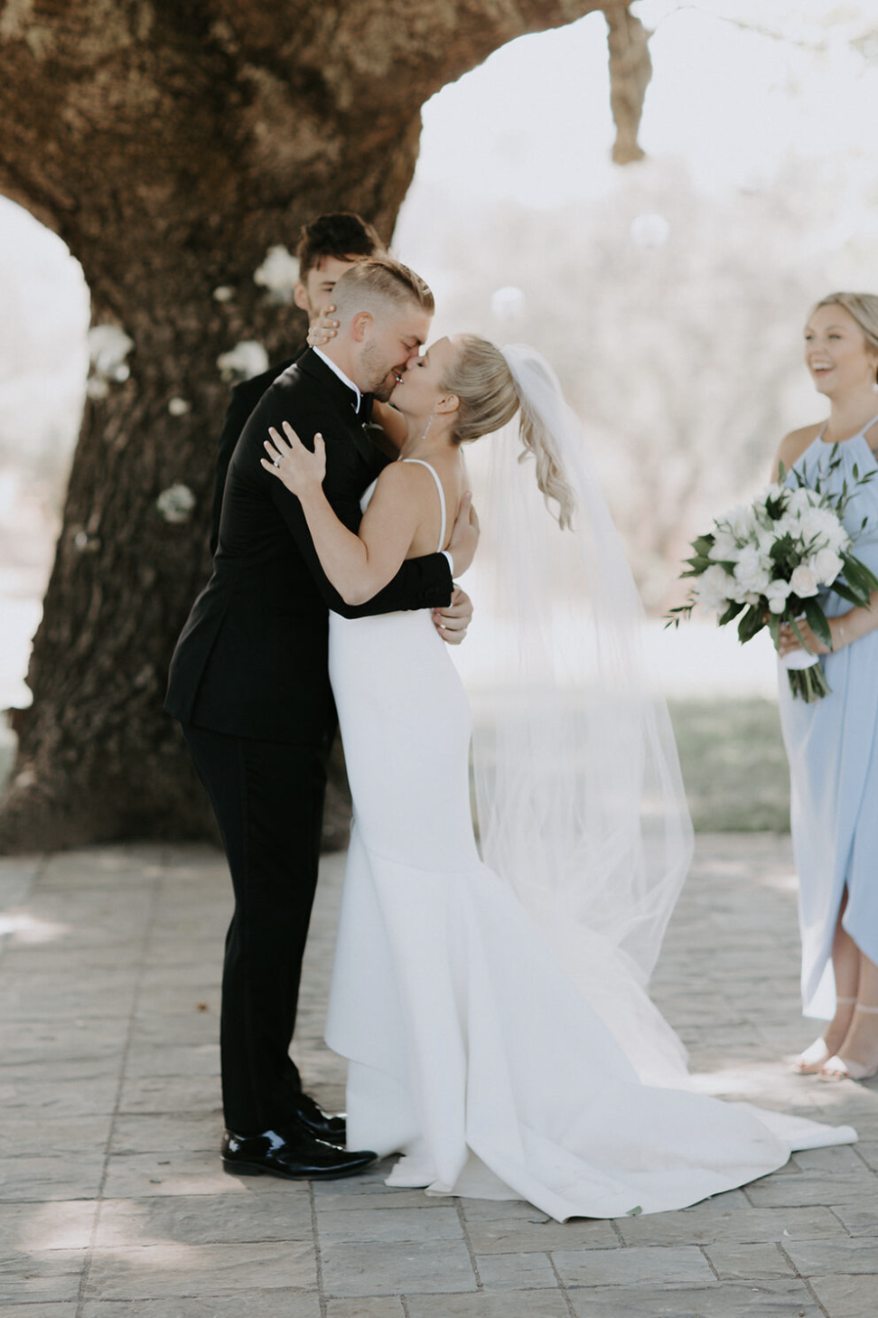 Bride and groom kissing under a large tree during ceremony, bridesmaid holding bouquet nearby.