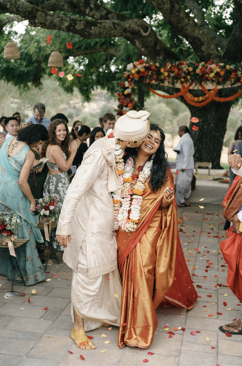 Couple in traditional Indian wedding attire celebrating joyfully under a decorated tree with guests.