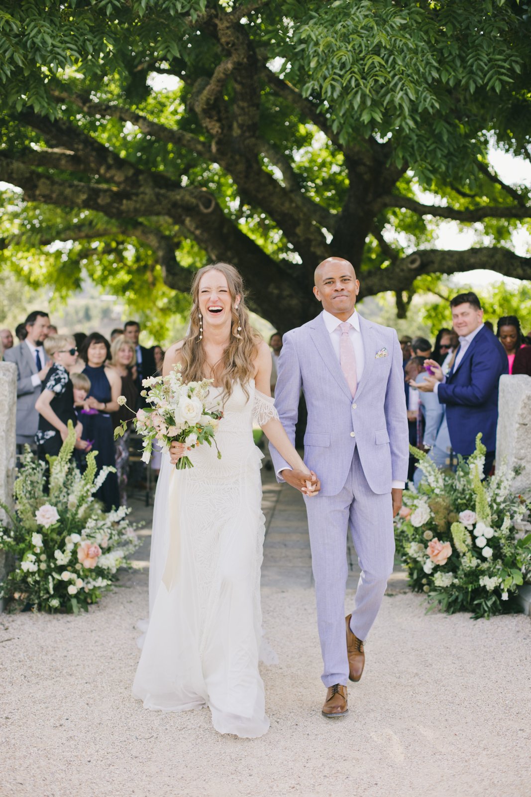 Bride and groom walking down the aisle after ceremony, guests cheering under a large tree decorated with flowers.