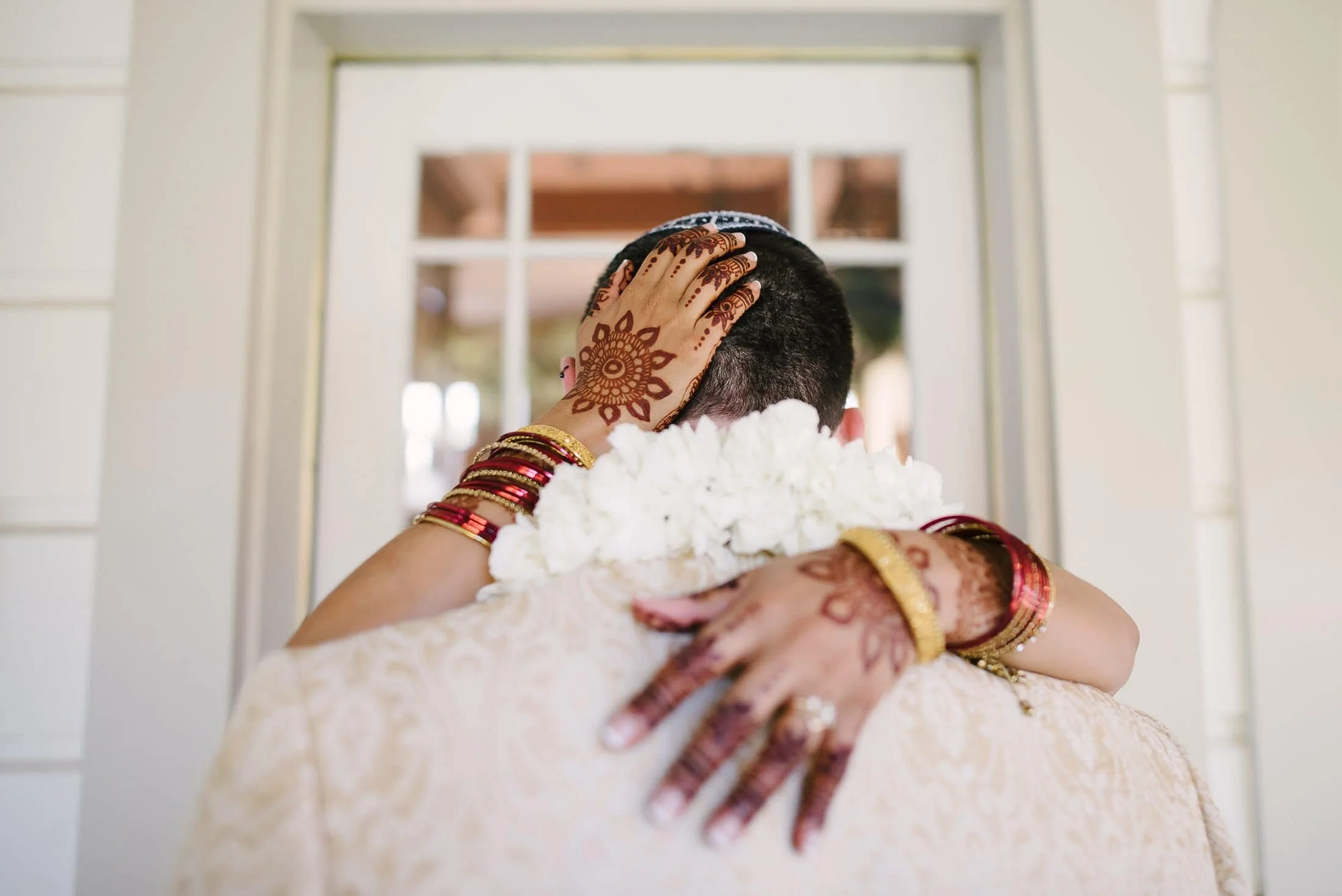 Bride in traditional Indian attire with jewelry and henna embracing partner in front of a mirror.