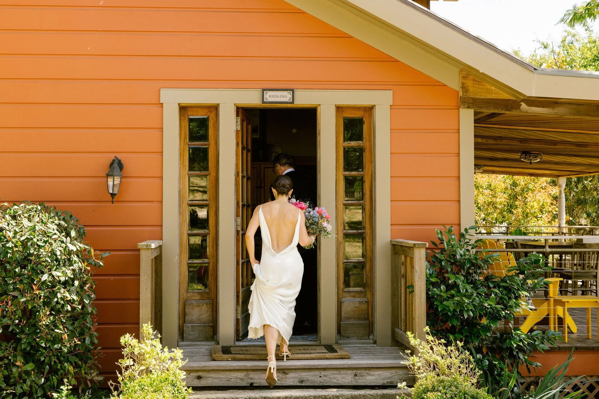 Bride in a white dress with an open back holding a bouquet, walking into a house at Triple S Ranch with a man inside, on a porch with plants and yellow chairs visible to the side.
