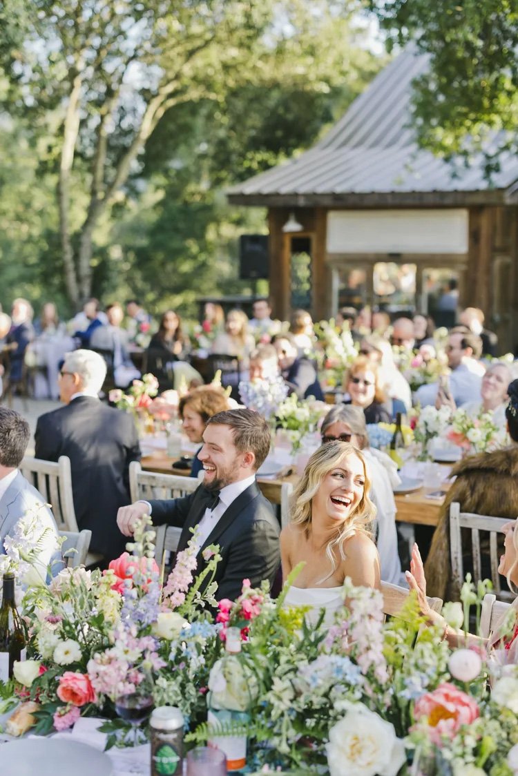 People at a wedding or outdoor celebration dinner at Triple S Ranch wedding venue in Napa, sitting at long tables decorated with colorful flowers, with a wooden building and green trees in the background.