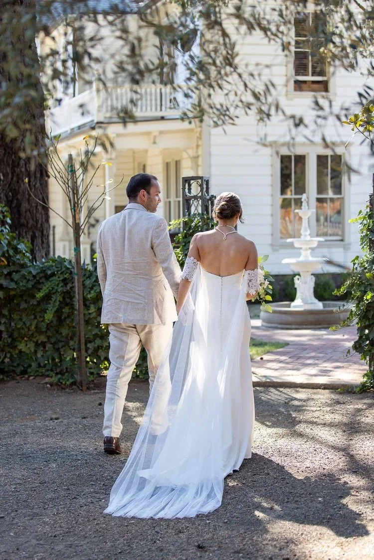 Bride and groom walking hand in hand in front of the Victorian Farmhouse at Triple S Ranch, with the bride wearing a white wedding dress and the groom in a light-colored suit.