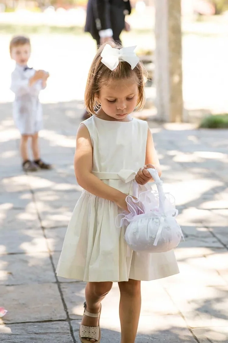 A wedding at Triple S Ranch in Napa with a young girl in a cream-colored dress with a large white bow in her hair holding a small white gift bag, walking outdoors on a paved pathway during daytime.
