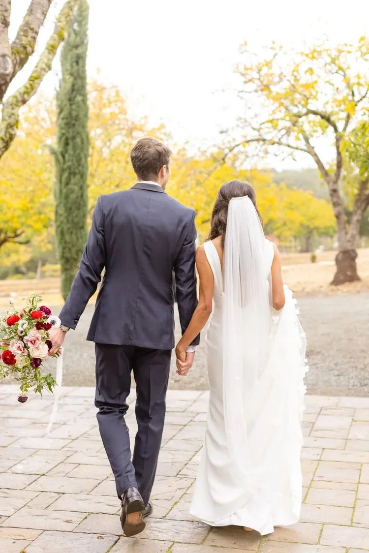 A bride and groom walking hand in hand outdoors at their Triple S Ranch Napa wedding. The bride is in a white wedding dress with a long veil, holding a bouquet of flowers, surrounded by autumn trees with yellow leaves.