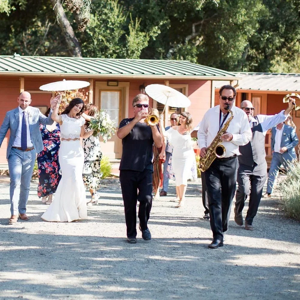 Group of people celebrating outdoors at a Napa wedding at Triple S Ranch, with a woman in a white dress holding a bouquet, and musicians playing saxophone and trumpet, under umbrellas, in front of a pink building with a green roof.