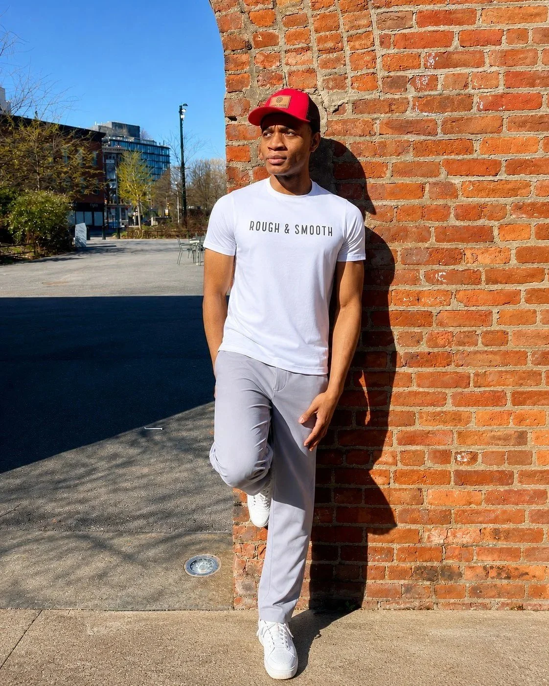 Male model wearing a Rough & Smooth white tee and red cap against a brick wall in New York.