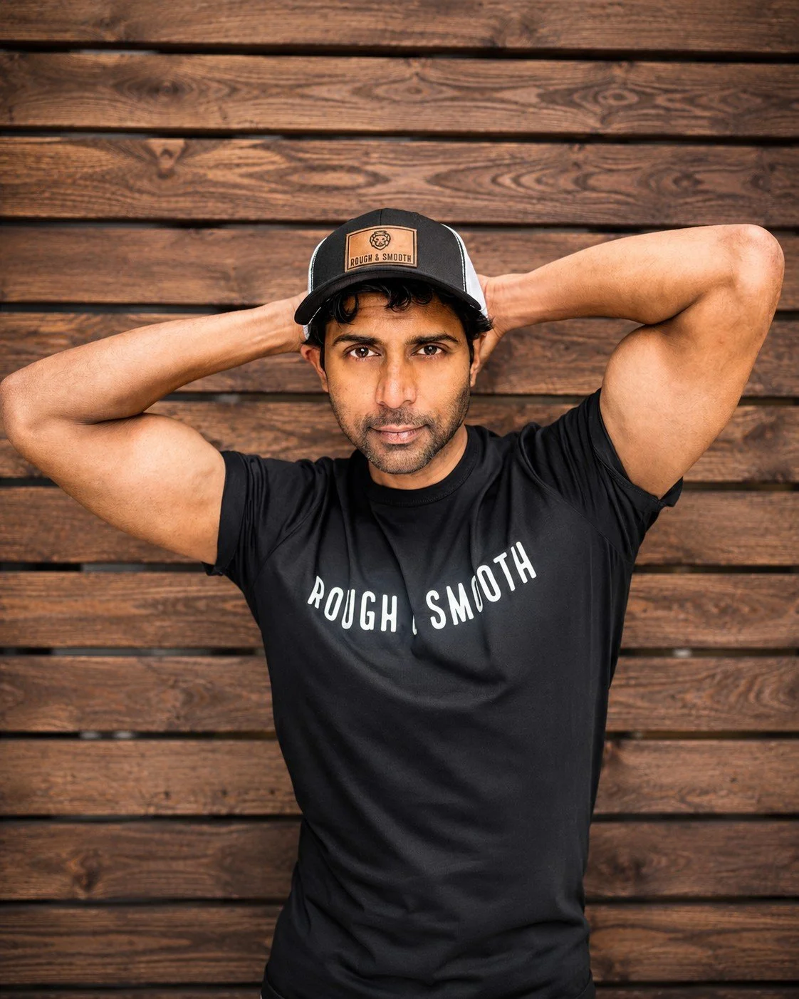 Male model wearing a Rough & Smooth black tee and signature cap in front of a wooden wall.