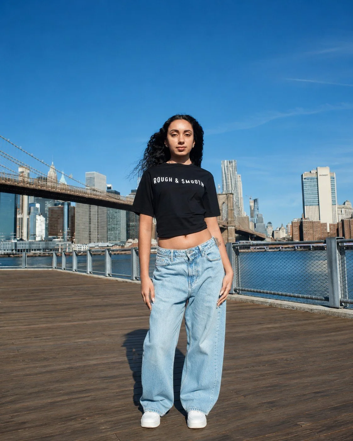 Female model wearing a Rough & Smooth black tee near the Brooklyn Bridge waterfront in New York.